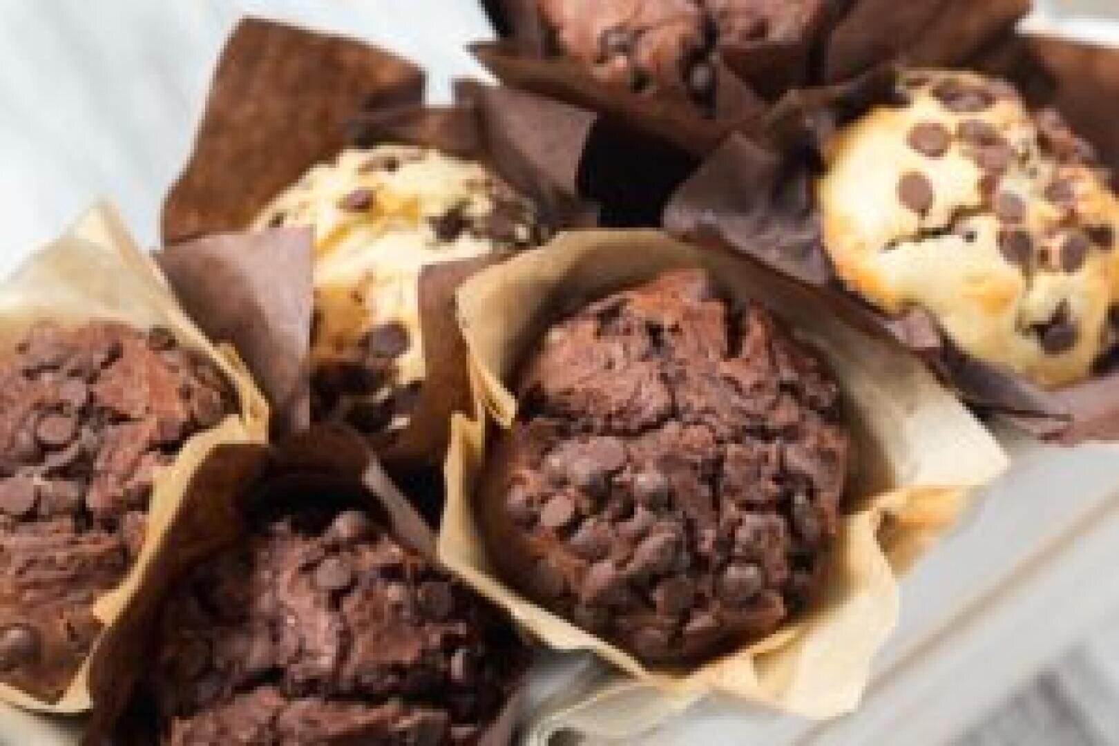 A close-up of assorted chocolate chip muffins, some chocolate and some vanilla, wrapped in brown and beige paper wrappers, arranged closely together on a white surface.