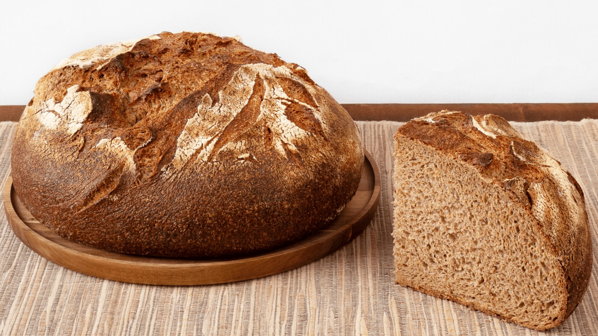Ein runder Laib rustikales Brot liegt neben einer einzelnen angeschnittenen Scheibe auf einem Holzteller. Das Ganze liegt auf einem beigefarbenen gewebten Tischset auf einem Holztisch mit weißem Hintergrund.