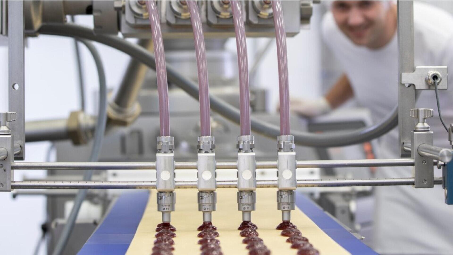 A close-up of a food production line, showing four nozzles dispensing red jelly onto rows of dough. In the background, a person in white work attire is smiling while monitoring the process.