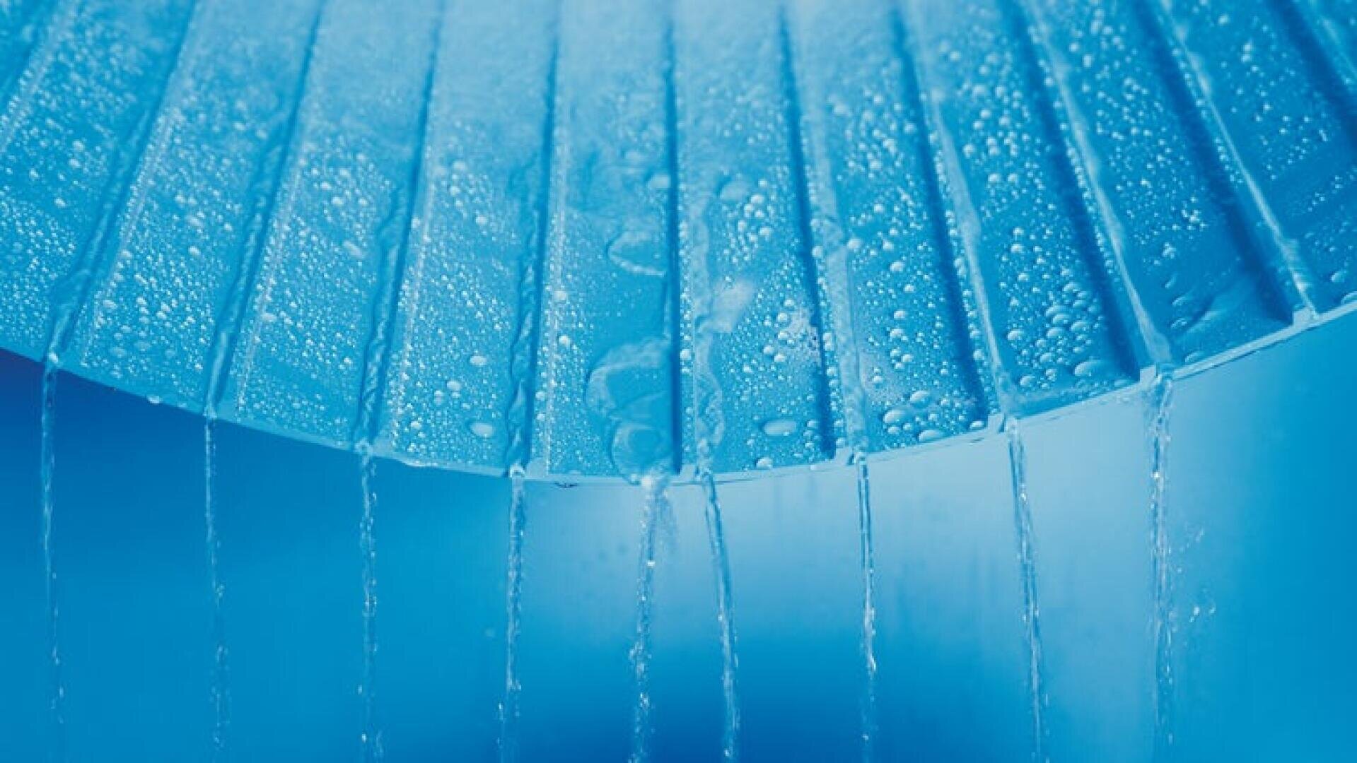 Nahaufnahme von Wassertropfen auf gekippten blauen Lamellen mit dünnen Wasserströmen, die zwischen den Lücken nach unten fließen, vor einem weichen blauen Hintergrund.