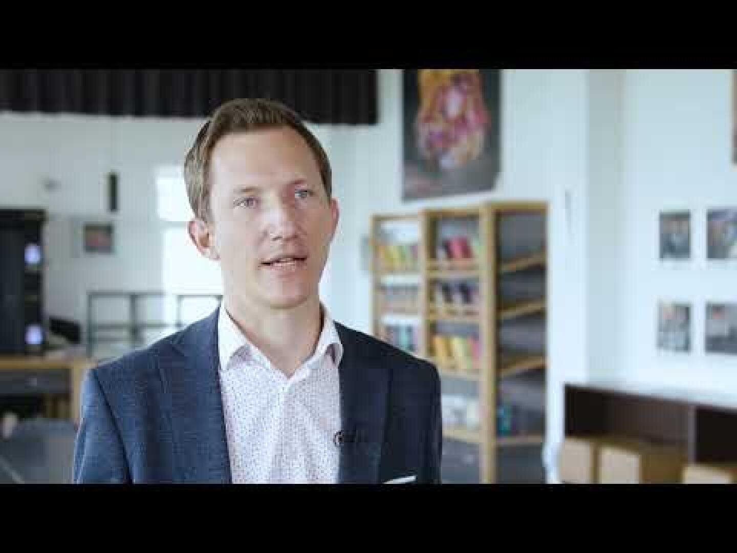 Ein Mann in einem blauen Blazer und einem weißen Hemd spricht in einem Innenbereich, möglicherweise in einem Klassenzimmer oder einer Bibliothek. Im Hintergrund sind Bücherregale und gerahmte Bilder zu sehen.