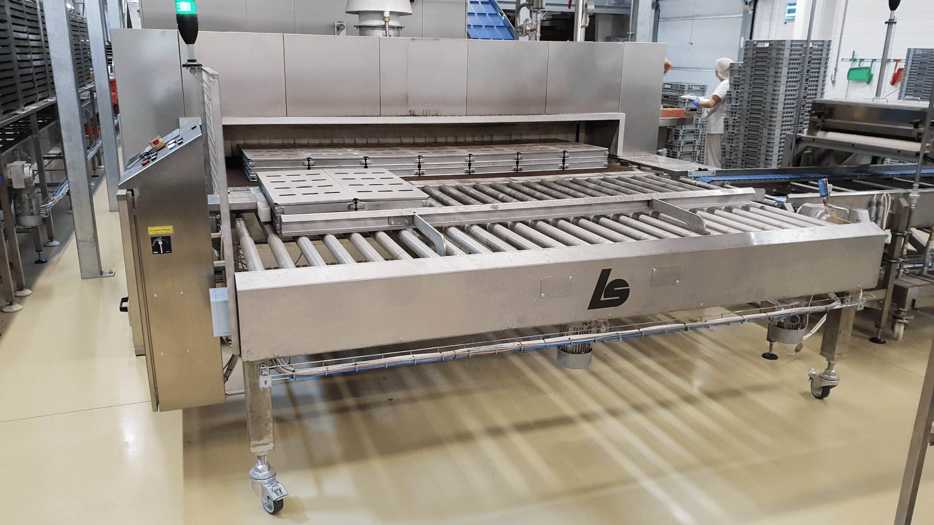 A large industrial stainless steel conveyor oven with rollers and control panel stands in a factory setting, surrounded by other equipment and metal racks on a clean, beige floor.