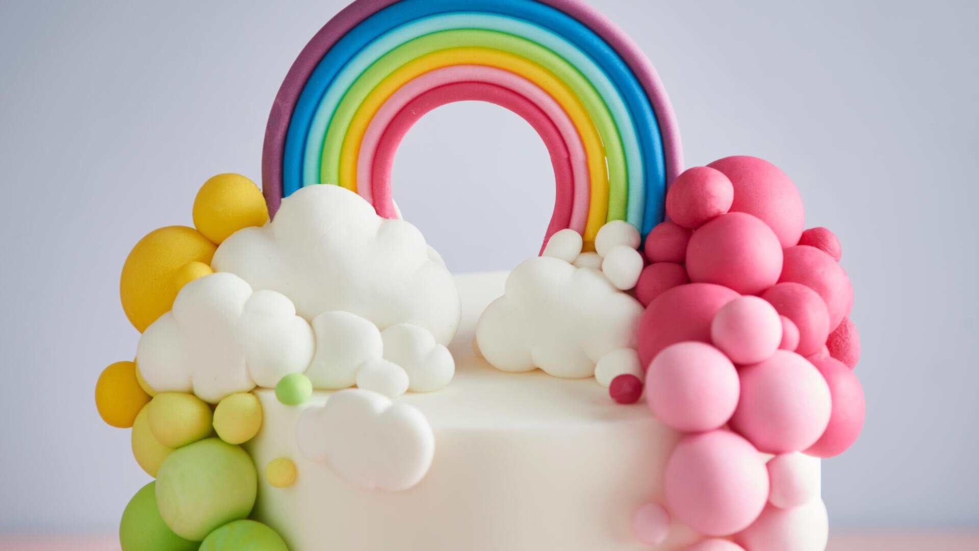 Ein Kuchen, dekoriert mit bunten Fondantblasen, weißen Wolken und einem Regenbogenbogen oben drauf. Die Blasen sind in Grün-, Gelb- und Rosatönen gehalten und ergeben ein skurril-fröhliches Design.