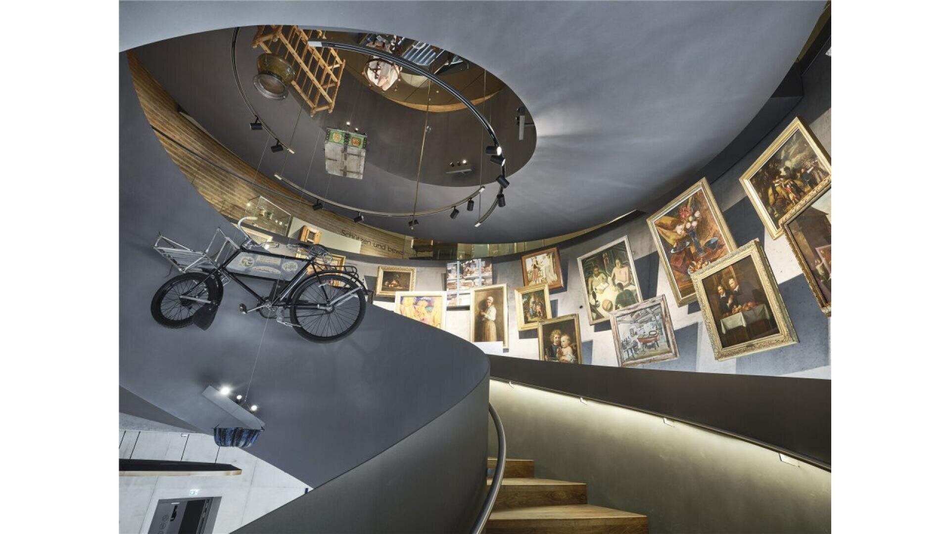 A spiral staircase leads upward beside a wall adorned with framed paintings; a vintage bicycle is mounted on the wall, and various objects are displayed above, creating an eclectic, museum-like atmosphere.