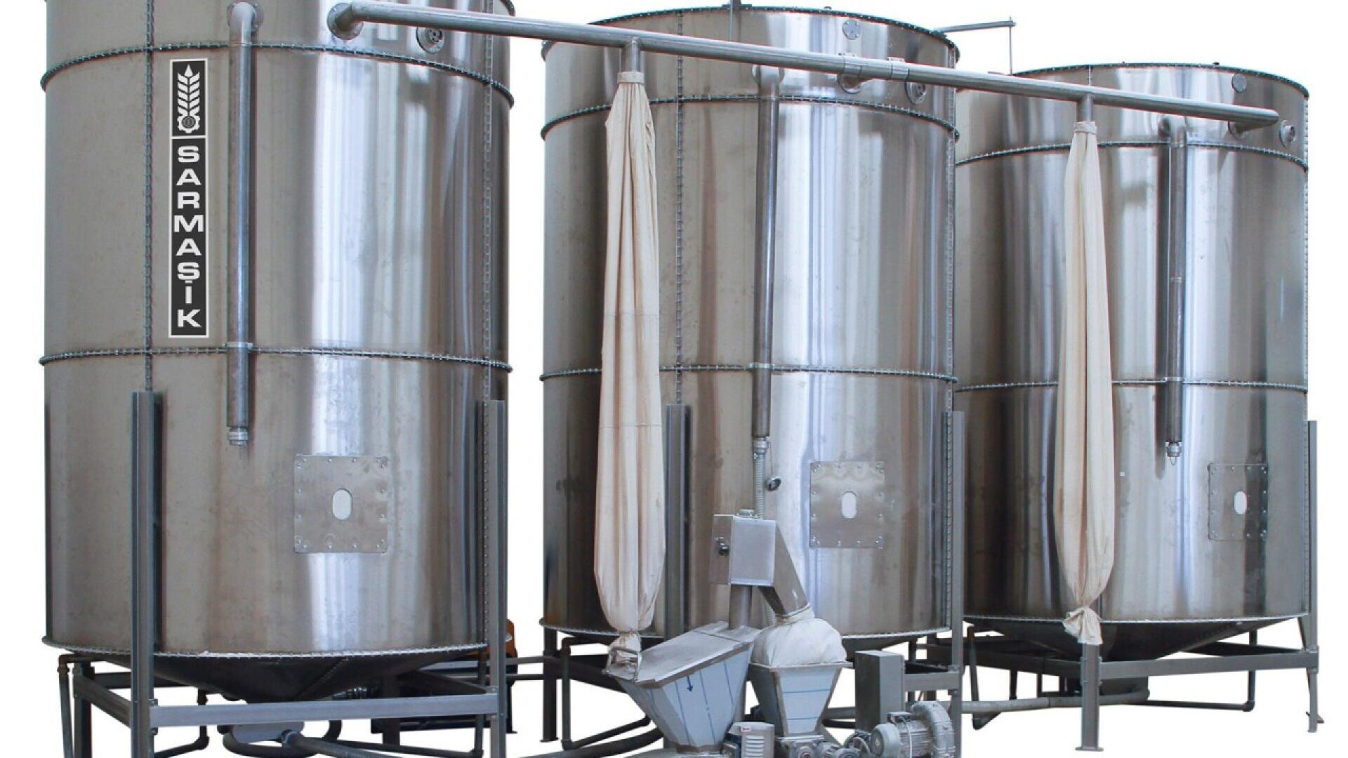 Three large, vertical stainless steel tanks with attached pipes and fabric filters stand in a row. The left tank is labeled “SARMAŞIK.” Industrial equipment is visible at the base of the tanks.