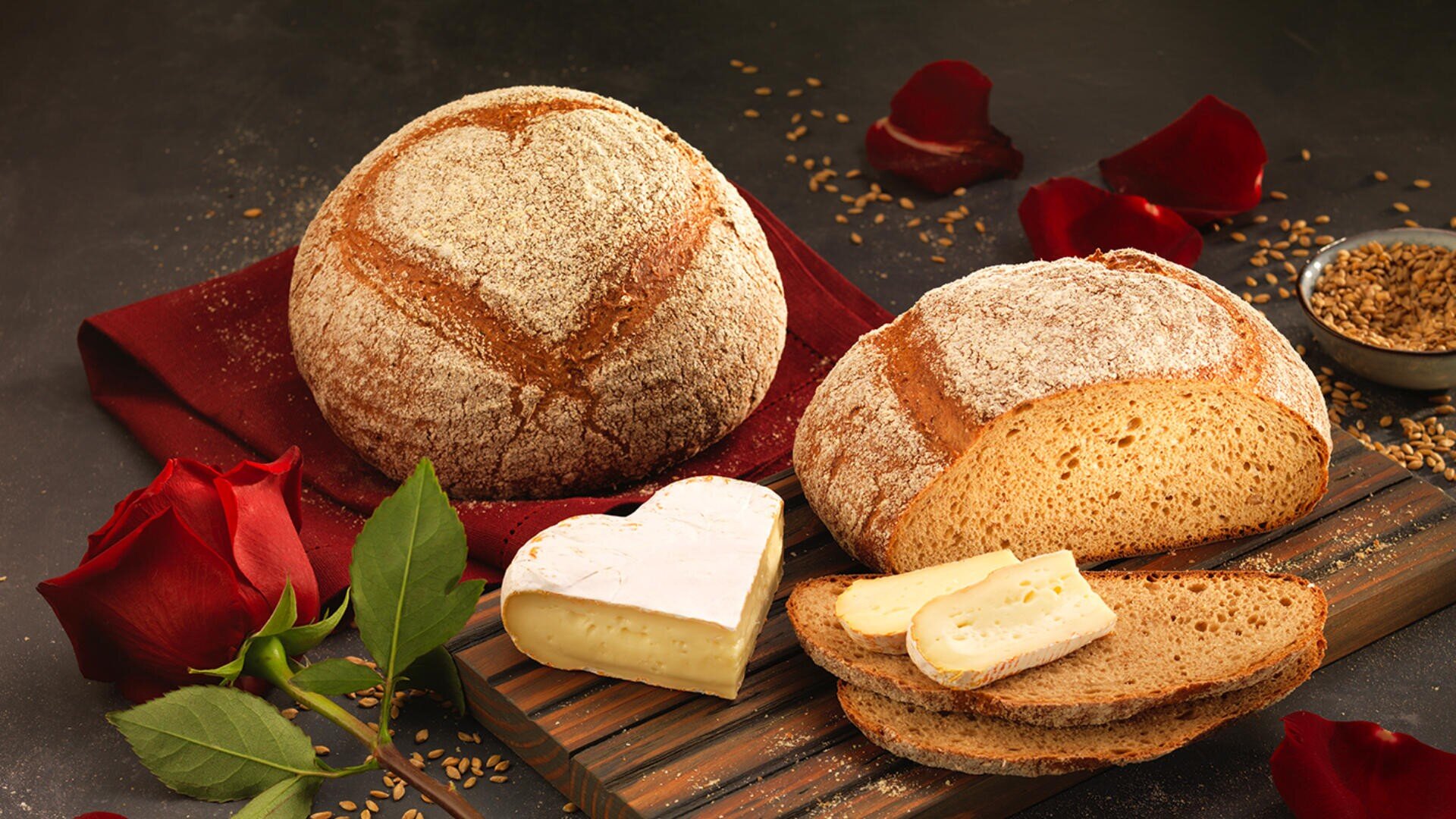 Zwei runde Laibe rustikales Brot, eines davon angeschnitten, liegen auf einem Holzbrett mit herzförmigen Käsescheiben. Rote Rosenblätter, eine ganze Rose und verstreute Samen verleihen dem dunklen, eleganten Ambiente eine romantische Note.