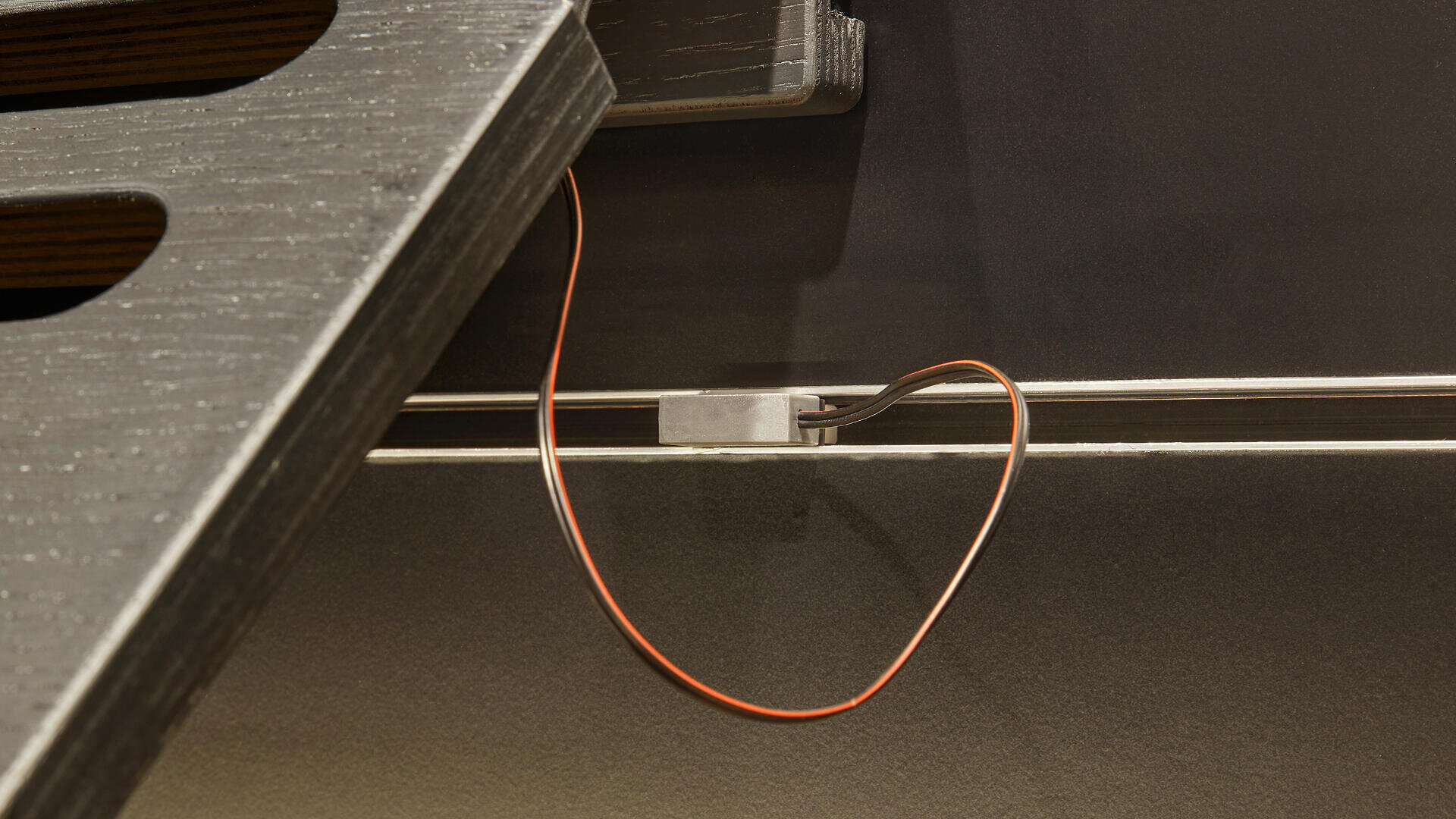 Nahaufnahme einer braunen Holztreppe mit einem dünnen orangefarbenen Draht, der mit einem kleinen Metallteil verbunden ist, das an der Wand unter der Treppe angebracht ist.