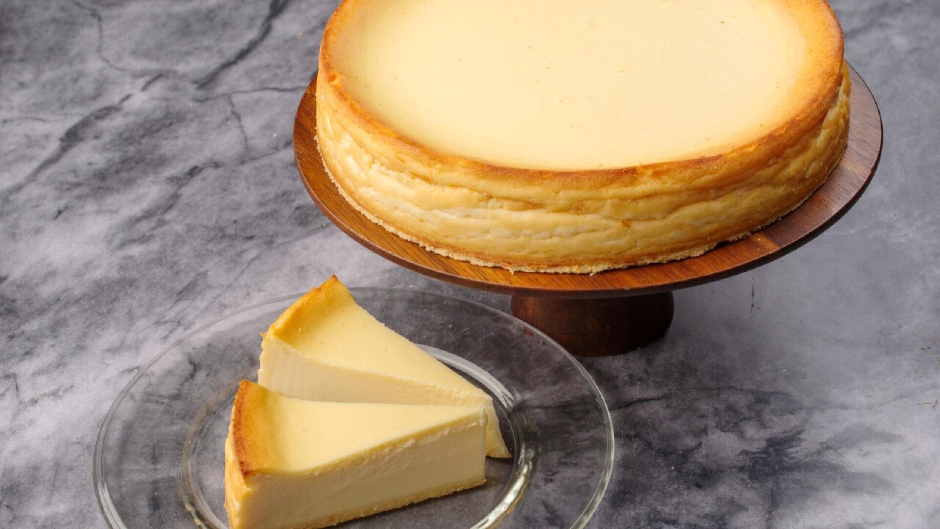 A whole cheesecake sits on a wooden cake stand, while two slices are placed on a clear glass plate nearby, all on a marble-textured surface.