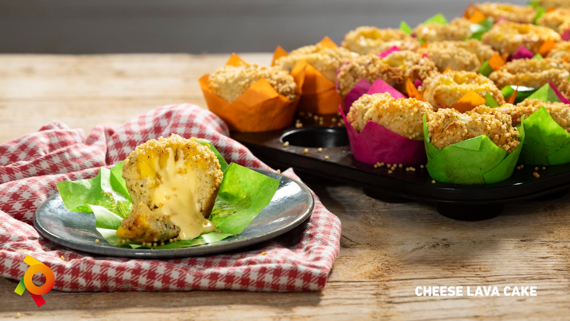 A tray of cheese lava cakes in colorful wrappers sits on a wooden table. One cake is cut open on a plate, showing melted cheese oozing out. A red-checked cloth and the text CHEESE LAVA CAKE are also visible.