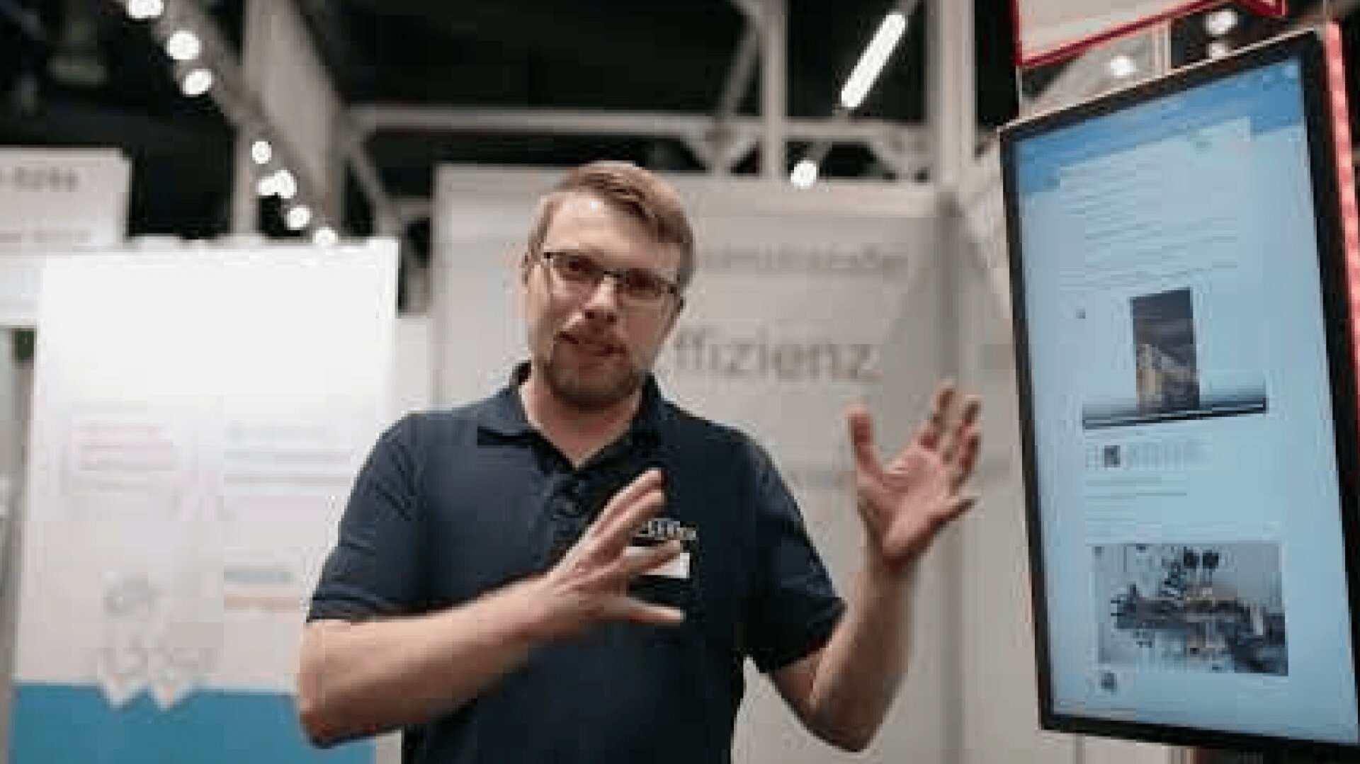 A man with glasses and a beard gestures with his hands while standing next to a large digital display screen at a modern indoor event or expo. Informational banners are visible in the background.