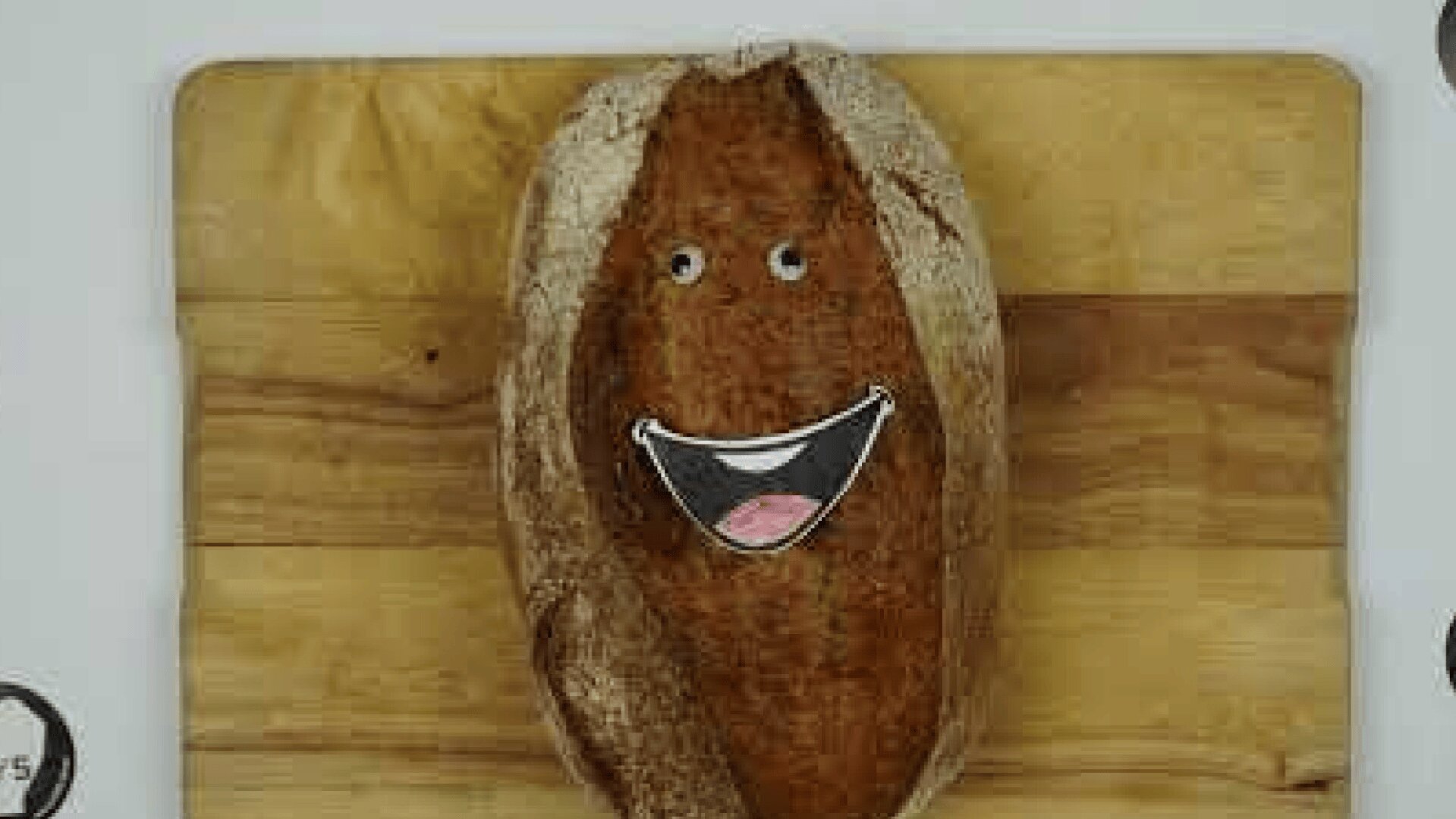 A loaf of bread on a wooden cutting board with cartoon eyes and a smiling mouth sticker, giving it a playful and happy face.
