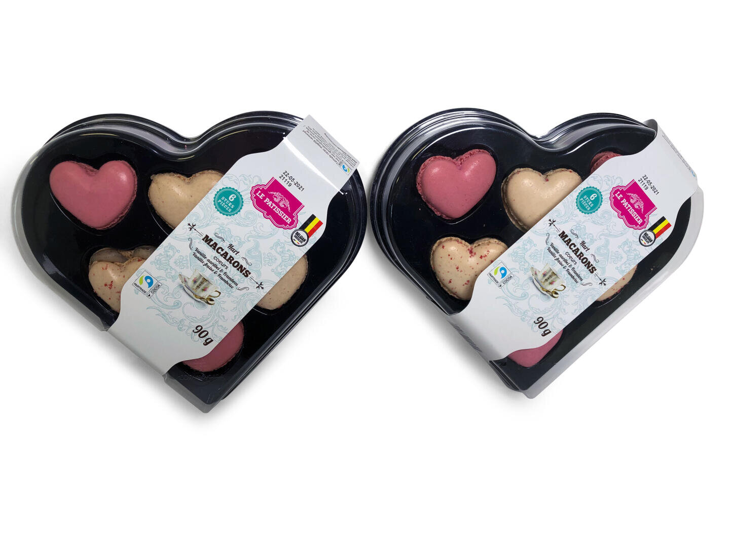 Zwei schwarze, herzförmige Schalen enthalten je vier herzförmige Macarons in Rosa und Beige, mit einer dekorativen Verpackung und einem Etikett mit Produktangaben und einer kleinen belgischen Flagge.