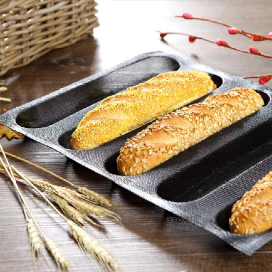A baking tray holds three baguettes topped with seeds, placed on a wooden table beside some wheat stalks, a wicker basket, and decorative red branches.