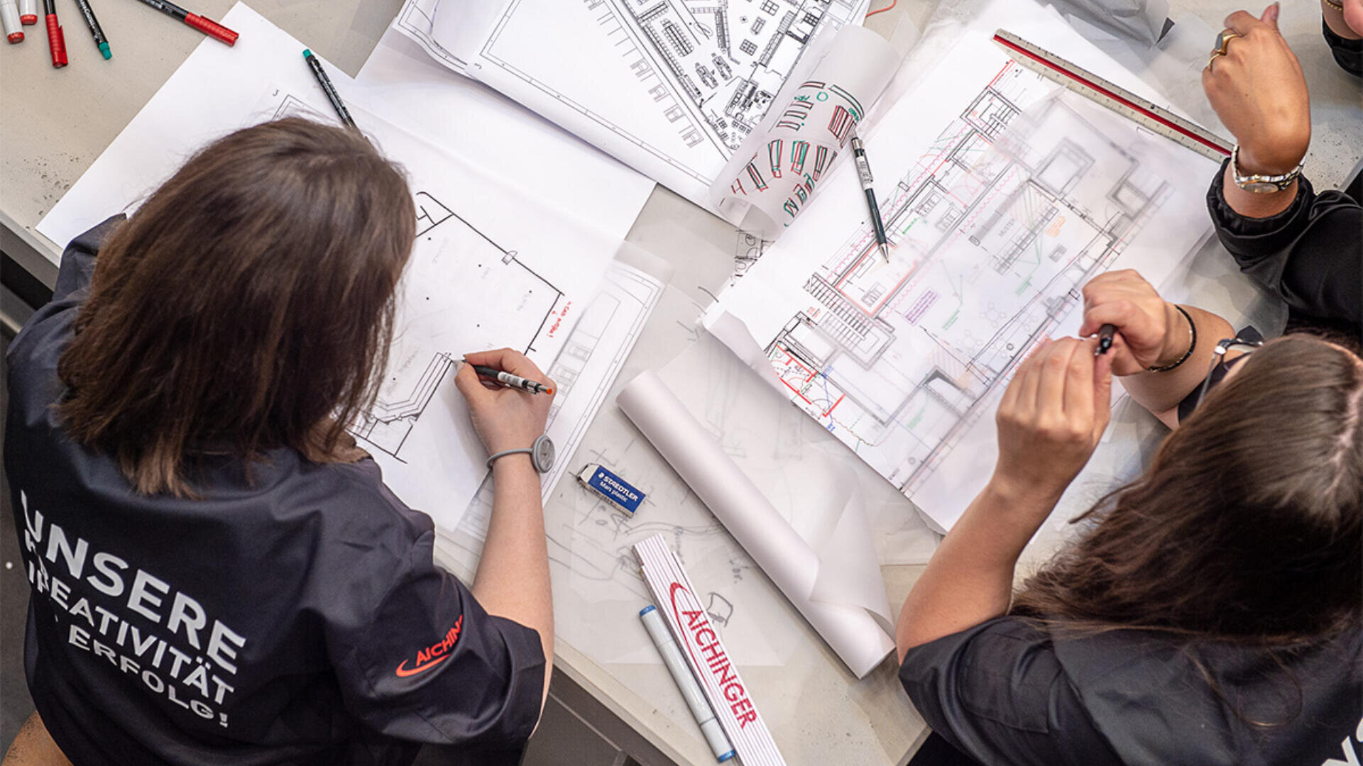 Zwei Personen arbeiten an einem Schreibtisch mit Stiften und Linealen an Architekturplänen. Verschiedene technische Zeichnungen, ein aufgerollter Plan und Schreibwaren liegen verstreut herum, und eine Person trägt ein schwarzes Hemd mit weißer Schrift auf dem Rücken.