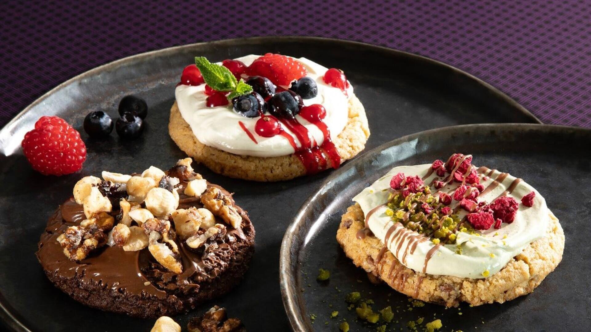 Three gourmet cookies on a dark plate: one topped with cream, berries, and mint; one with chocolate spread, nuts, and caramel; and one with cream, pistachios, and dried raspberries.