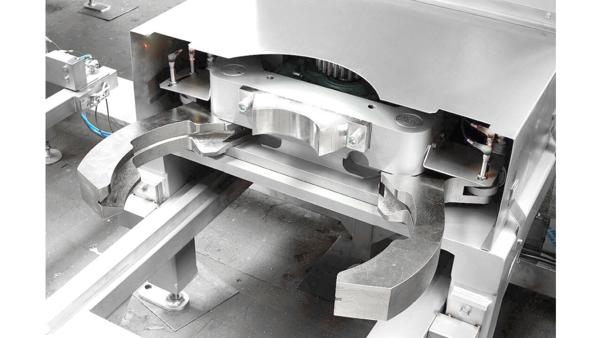 Nahaufnahme einer großen, industriellen Metallmaschine mit gebogenen, klauenartigen Armen und polierten Oberflächen, die über Metallschienen in einer Fabrik oder Werkstatt positioniert ist.