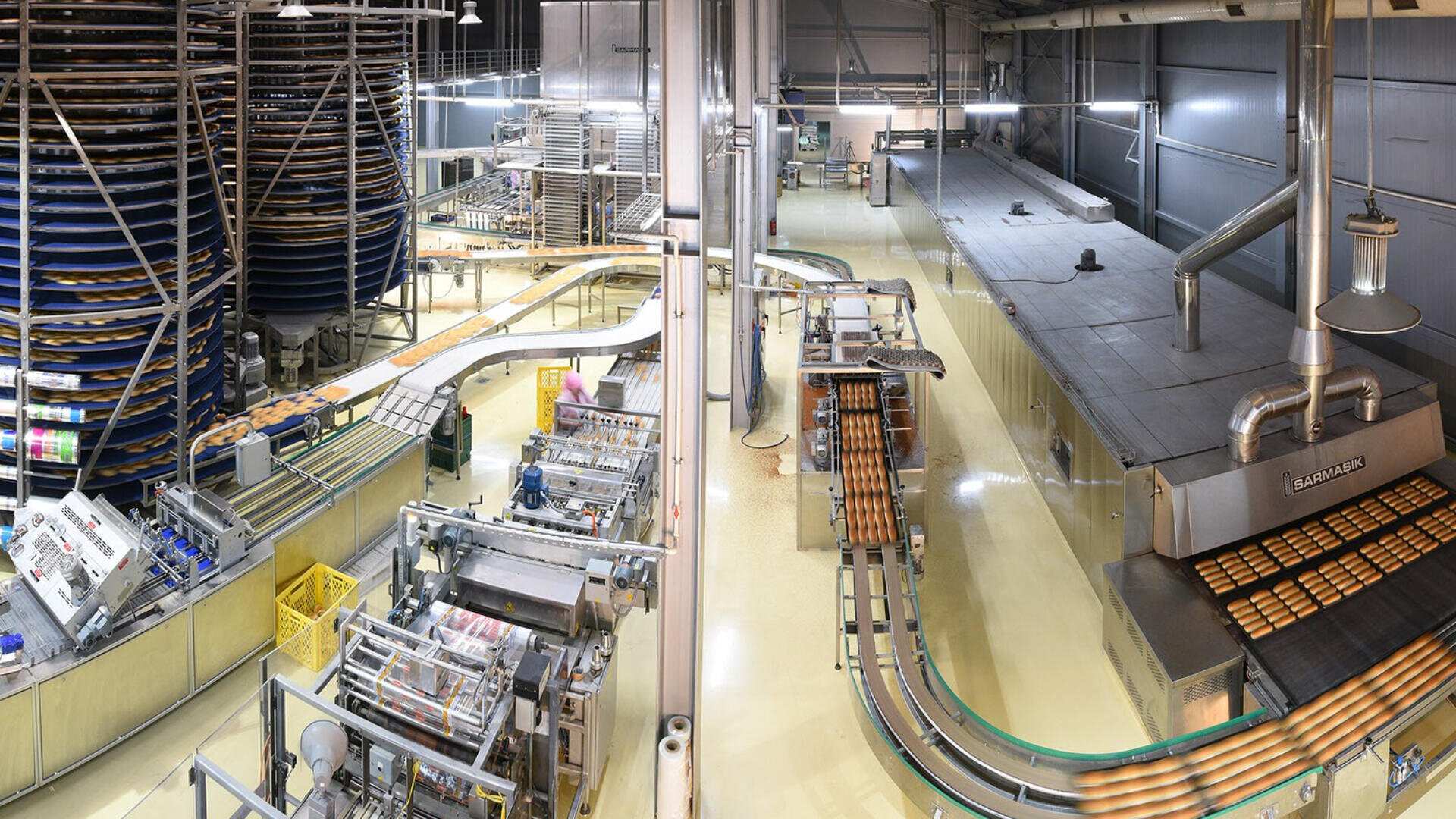 A modern industrial bakery with automated machines, conveyor belts, and large ovens producing and packaging bread loaves in a clean, brightly lit facility.