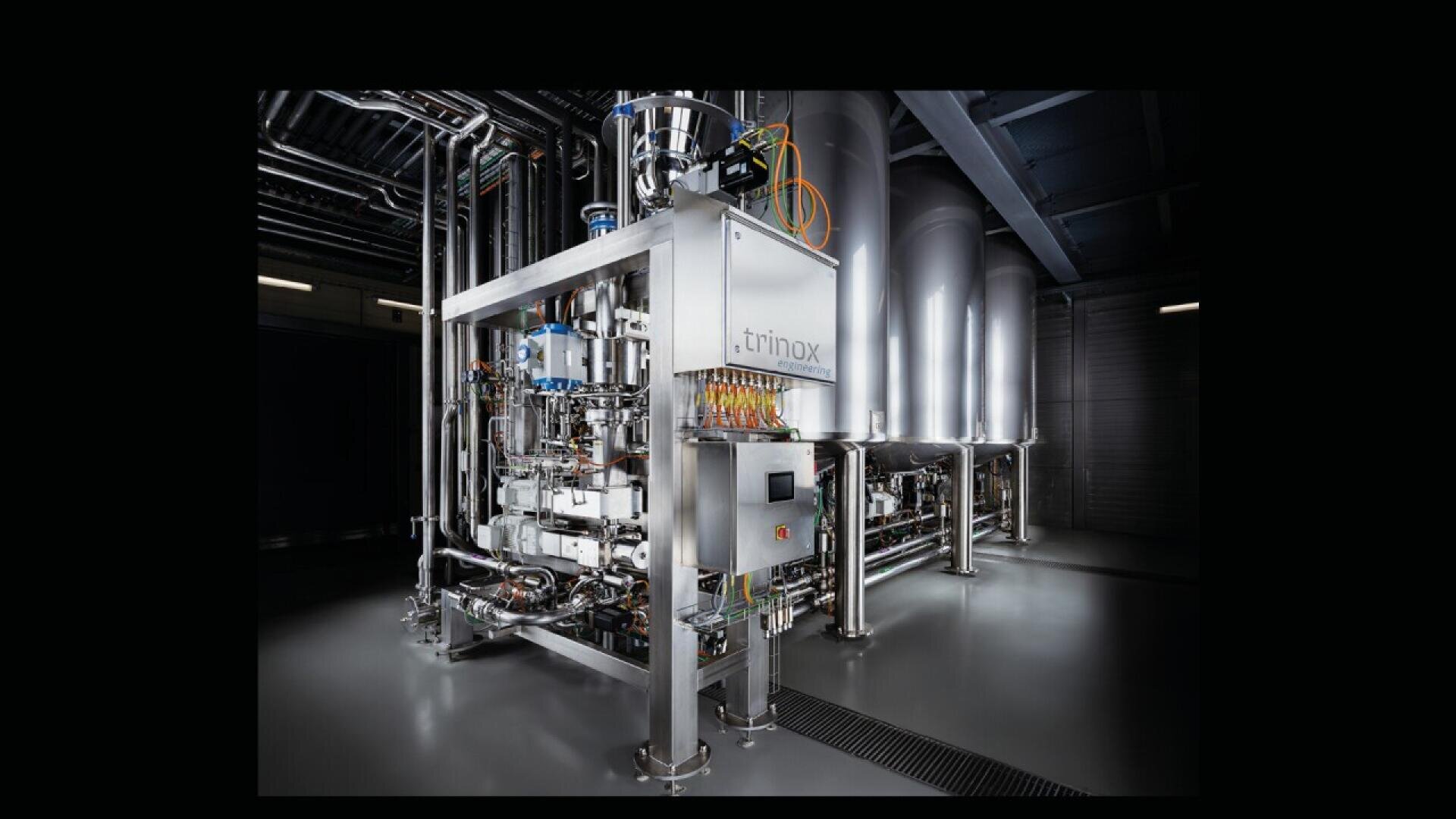 A large, stainless steel industrial machine with pipes, wires, and control panels stands in a dimly lit factory setting, with tall cylindrical tanks in the background.