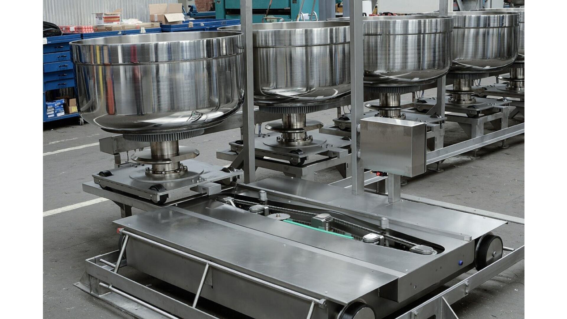 Mehrere große industrielle Mischmaschinen aus rostfreiem Stahl sind in einer Fabrik- oder Werkstatteinrichtung in einer Reihe aufgestellt, umgeben von verschiedenen Geräten und blauen Lagerschubladen.