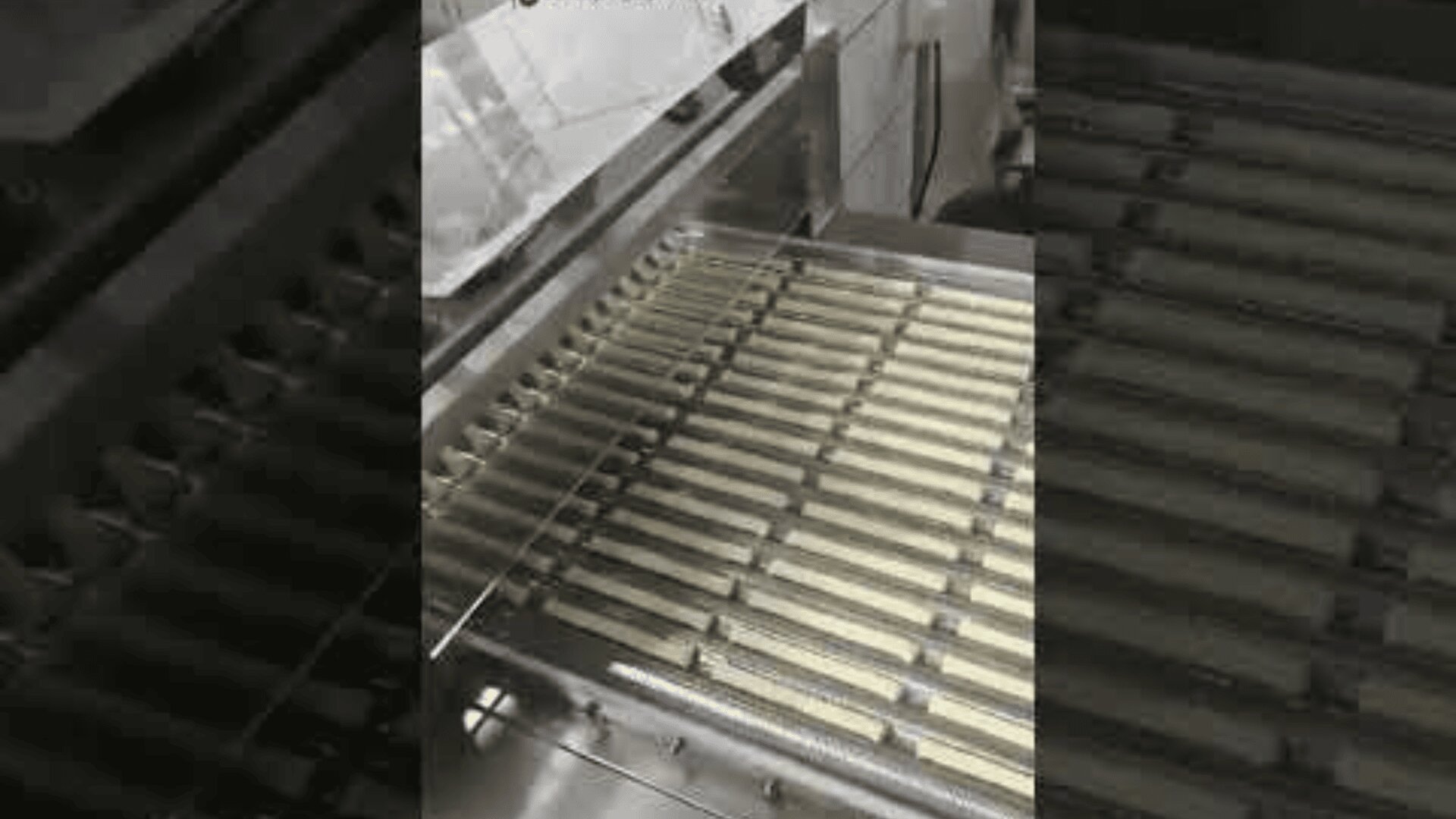 Ein Förderband in einer Fabrik transportiert Reihen von rechteckigen Keksen unter einem Metallgitter in einer industriellen Lebensmittelverarbeitungsmaschine.