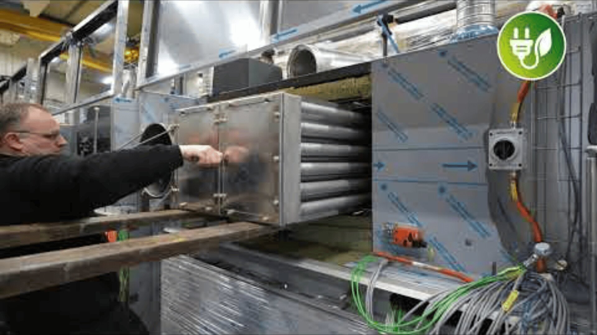 Eine Person arbeitet an einer Industriemaschine mit Metallrohren und Kabeln. Das Äußere der Maschine ist silbern mit blauen Pfeilen, und in der oberen rechten Ecke ist ein Symbol für Energieeffizienz zu sehen.