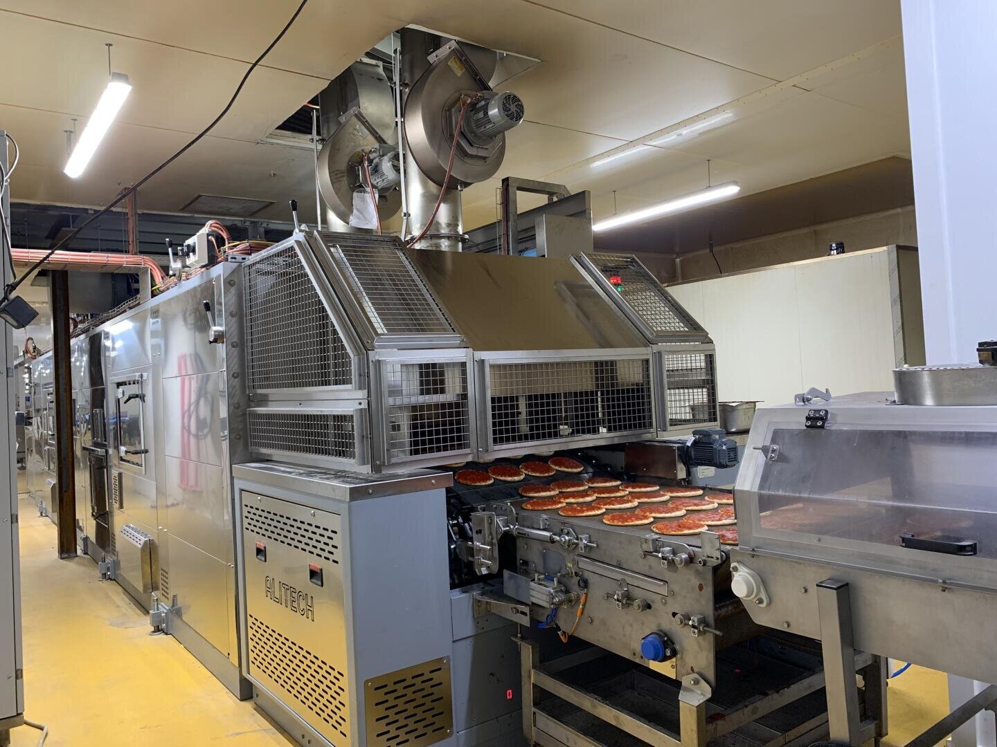 A large industrial pizza oven conveyor with multiple pizzas moving through the machine in a food processing factory, surrounded by metal equipment and bright lighting.