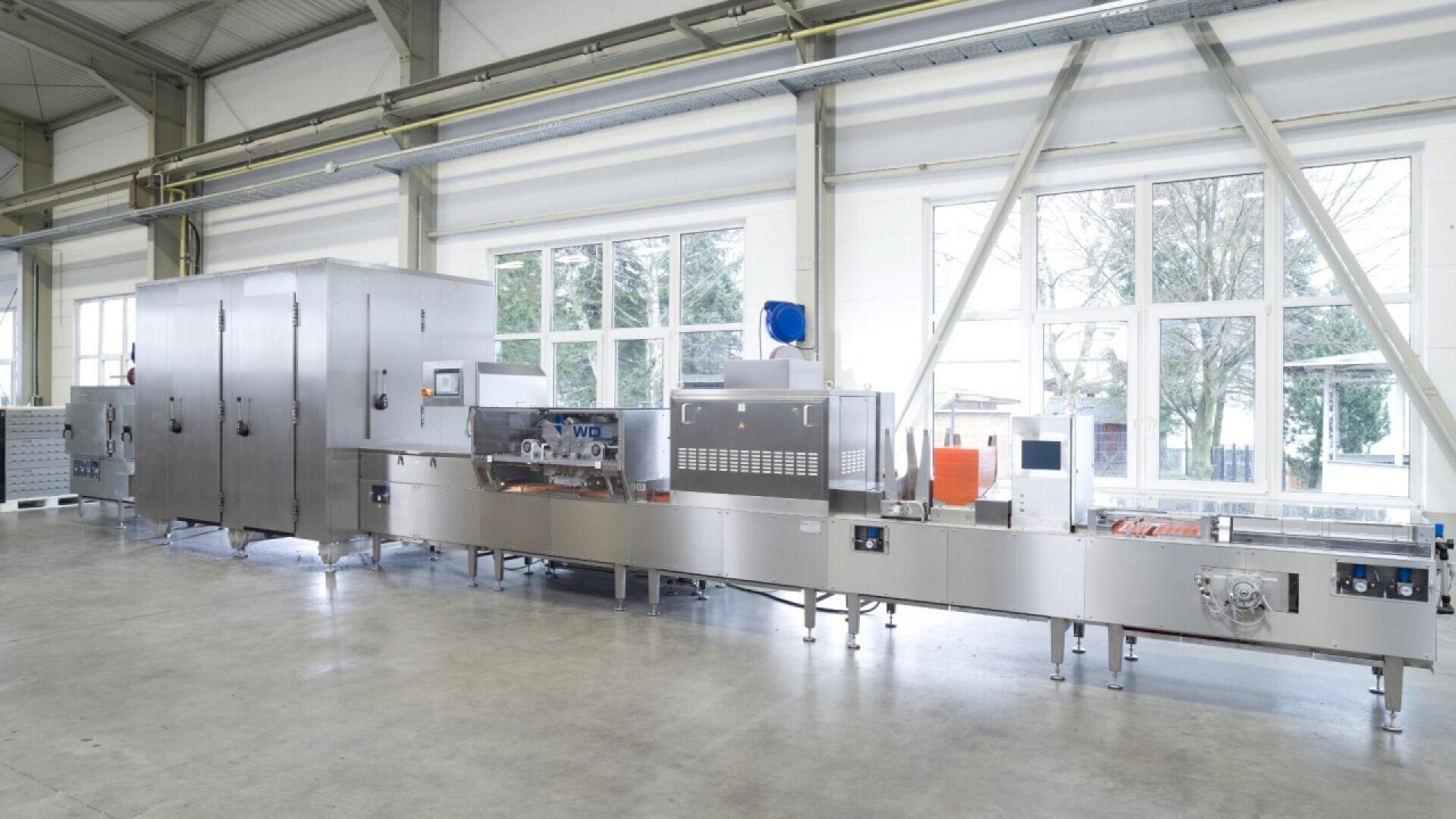 A large industrial food processing machine with stainless steel components stands in a bright, spacious factory with large windows showing trees outside. The equipment has control panels and conveyor belts.