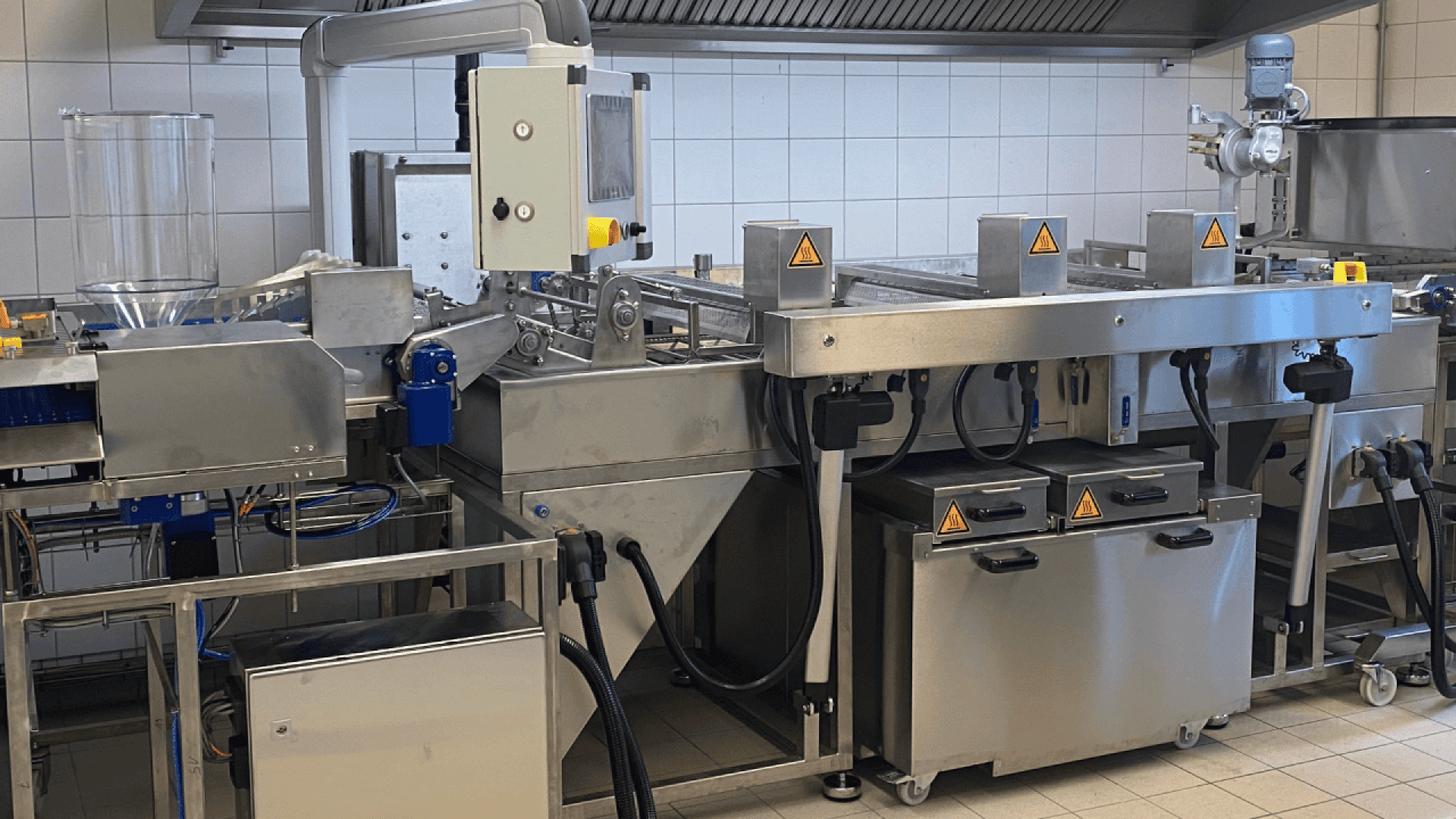 A large stainless steel industrial food processing machine with control panels and safety warning signs, set in a clean, tiled commercial kitchen environment.