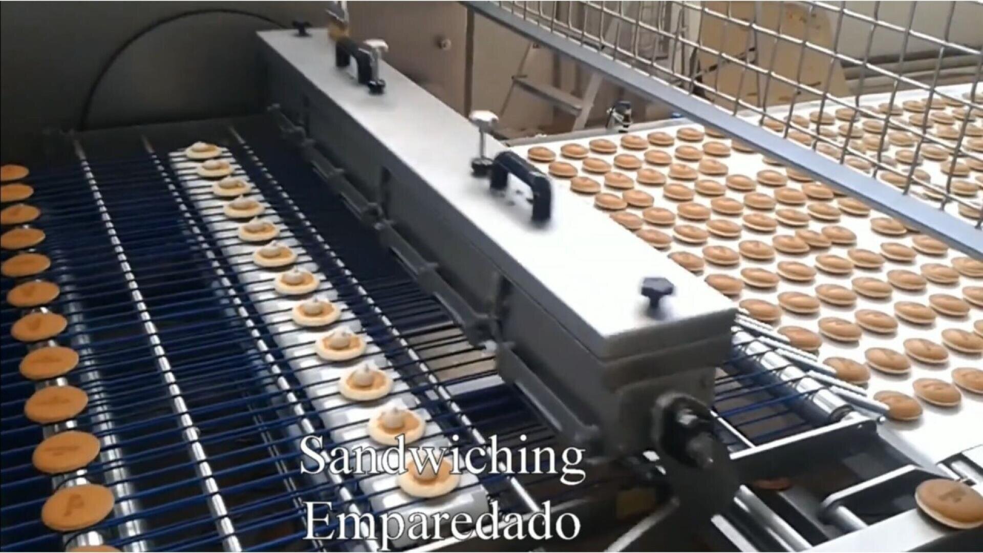 Eine Fabrikmaschine schichtet automatisch Sahne zwischen Reihen von runden Keksen auf einem Förderband ein. Der Text Sandwiching Emparedado erscheint am unteren Rand des Bildes.