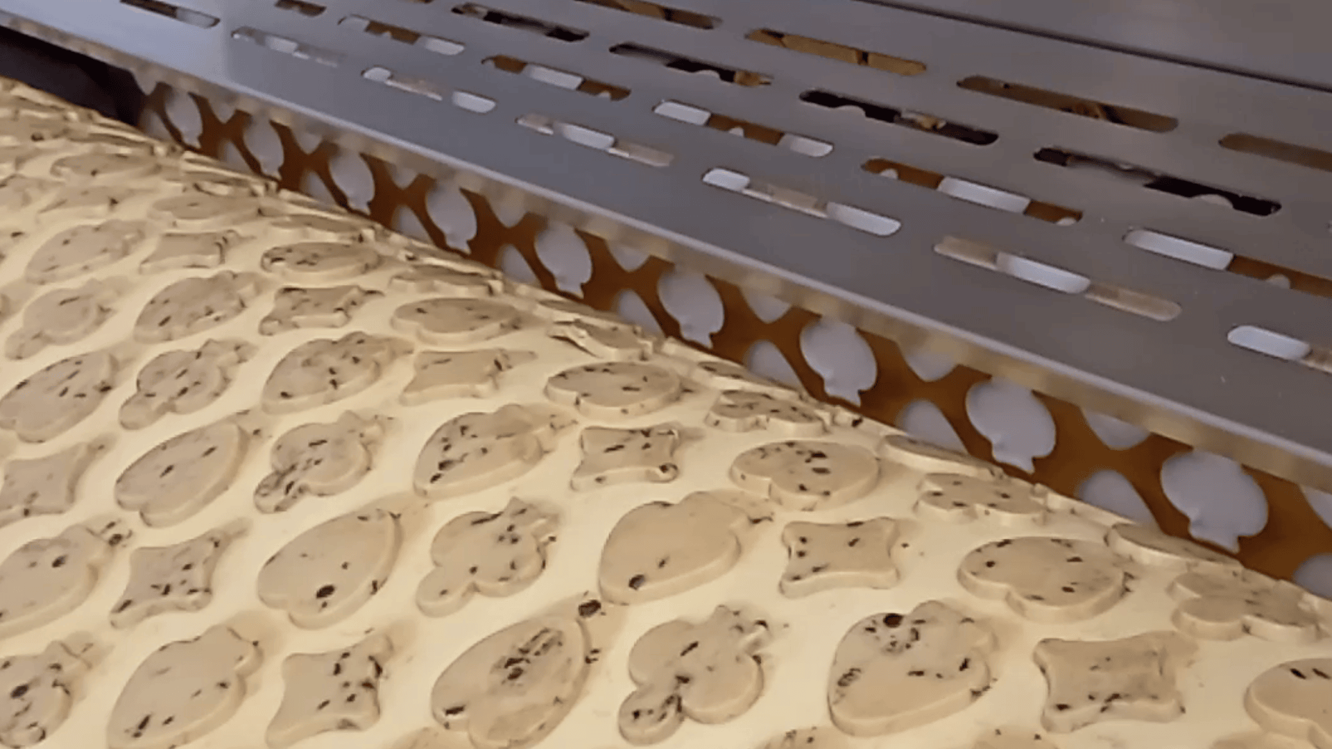 A machine presses shapes into a sheet of cookie dough embedded with chocolate chips, forming rows of animal faces, likely part of an automated baking process.