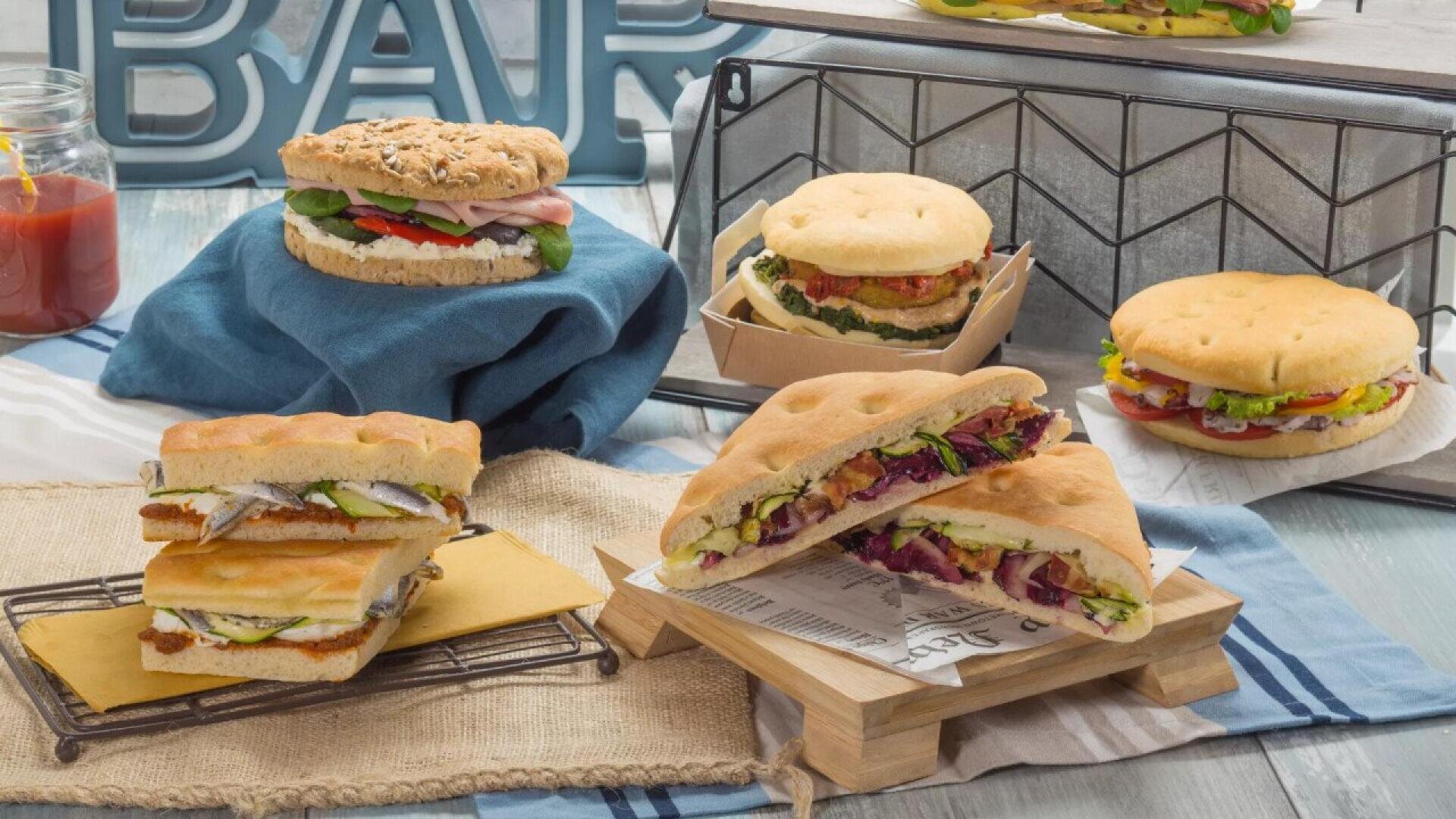 A variety of sandwiches with vegetables, cheese, and deli meats displayed on trays and baskets atop a decorated table with a jar of ketchup and a large BAR sign in the background.