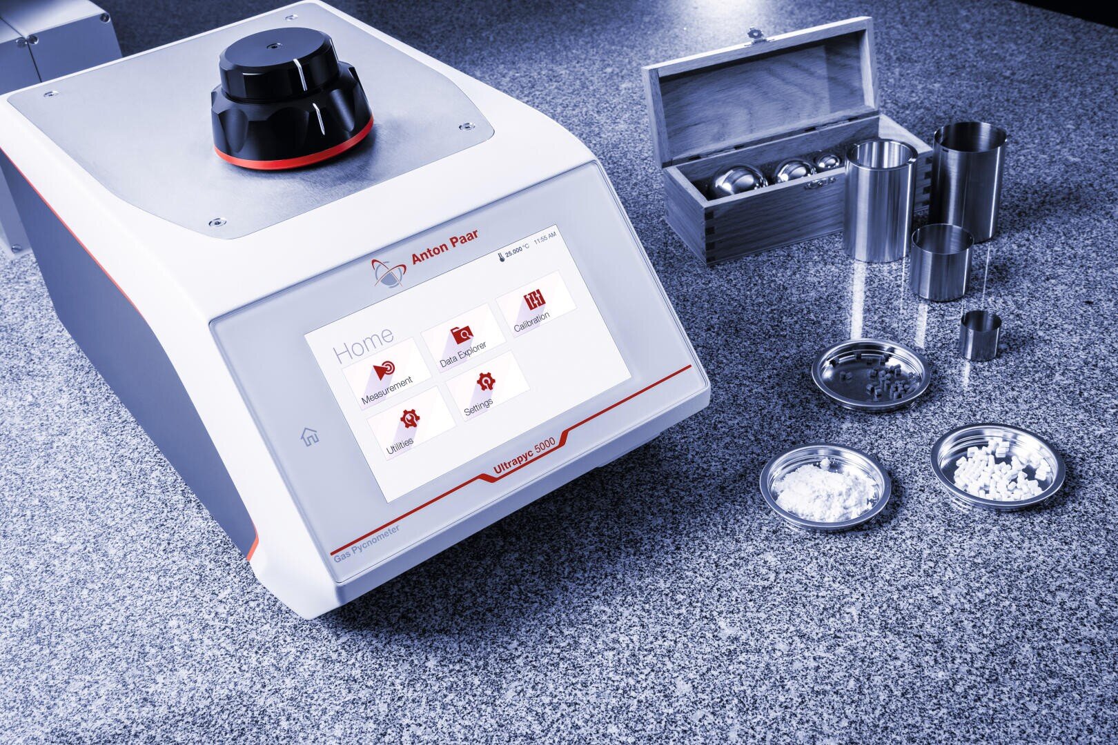 A laboratory grinder with a digital touchscreen interface sits on a countertop next to metal grinding containers, powdered samples in dishes, and a wooden box holding metal balls.