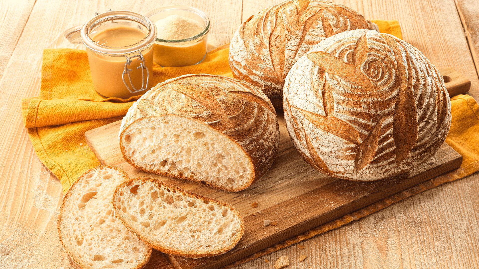 Round loaves of rustic bread, one sliced to show the airy crumb, sit on a wooden board with two slices in front. A jar of honey and a bowl of flour are nearby on a yellow cloth atop a wooden table.