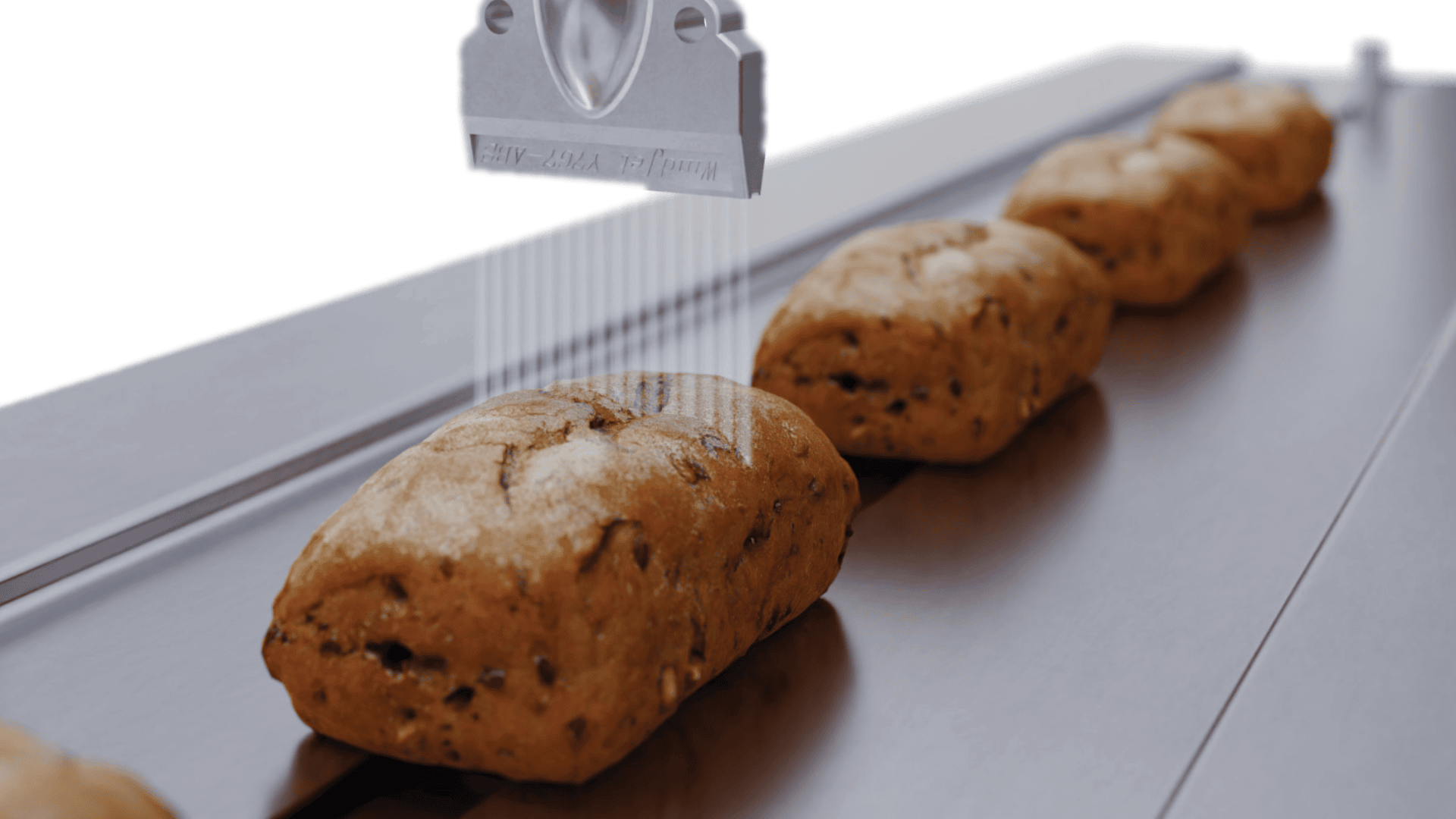 Eine Nahaufnahme von Brotlaiben, die sich auf einem Förderband bewegen, mit einer Maschine darüber, die etwas gleichmäßig auf das Brot sprüht oder aufträgt.