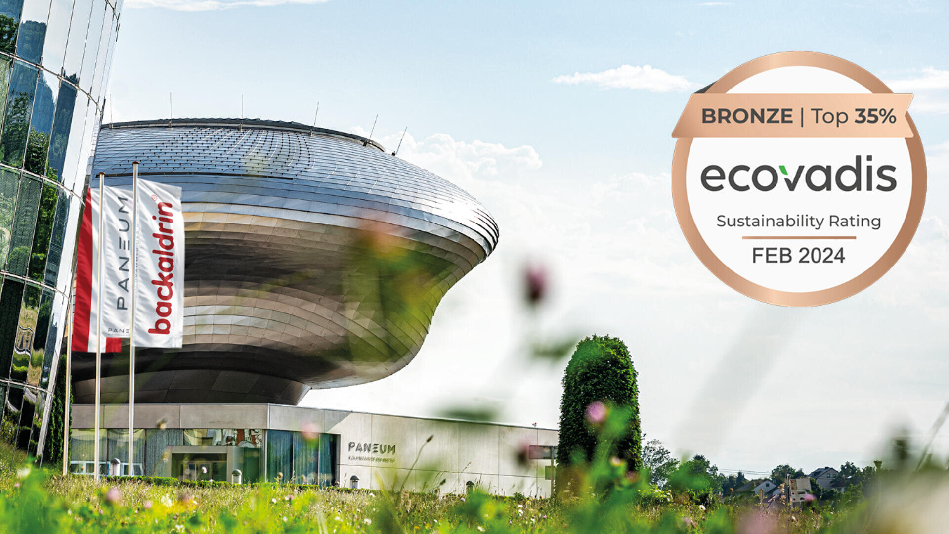 Ein modernes, silbernes Gebäude mit geschwungener Architektur steht hinter zwei Fahnen mit der Aufschrift PANEUM und backaldrin. Im Vordergrund ist eine bronzene Ecovadis-Plakette für die Nachhaltigkeitsbewertung im Februar 2024 zu sehen.