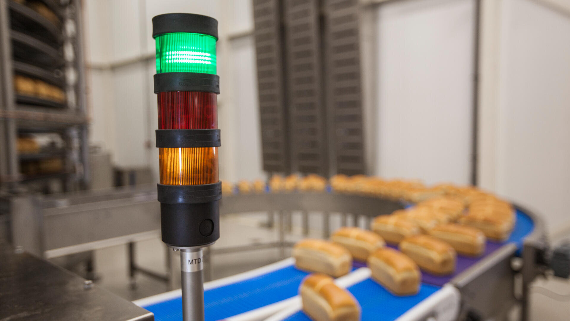 A green, yellow, and red signal light stands near a conveyor belt carrying rows of baked bread loaves in a modern food processing facility.