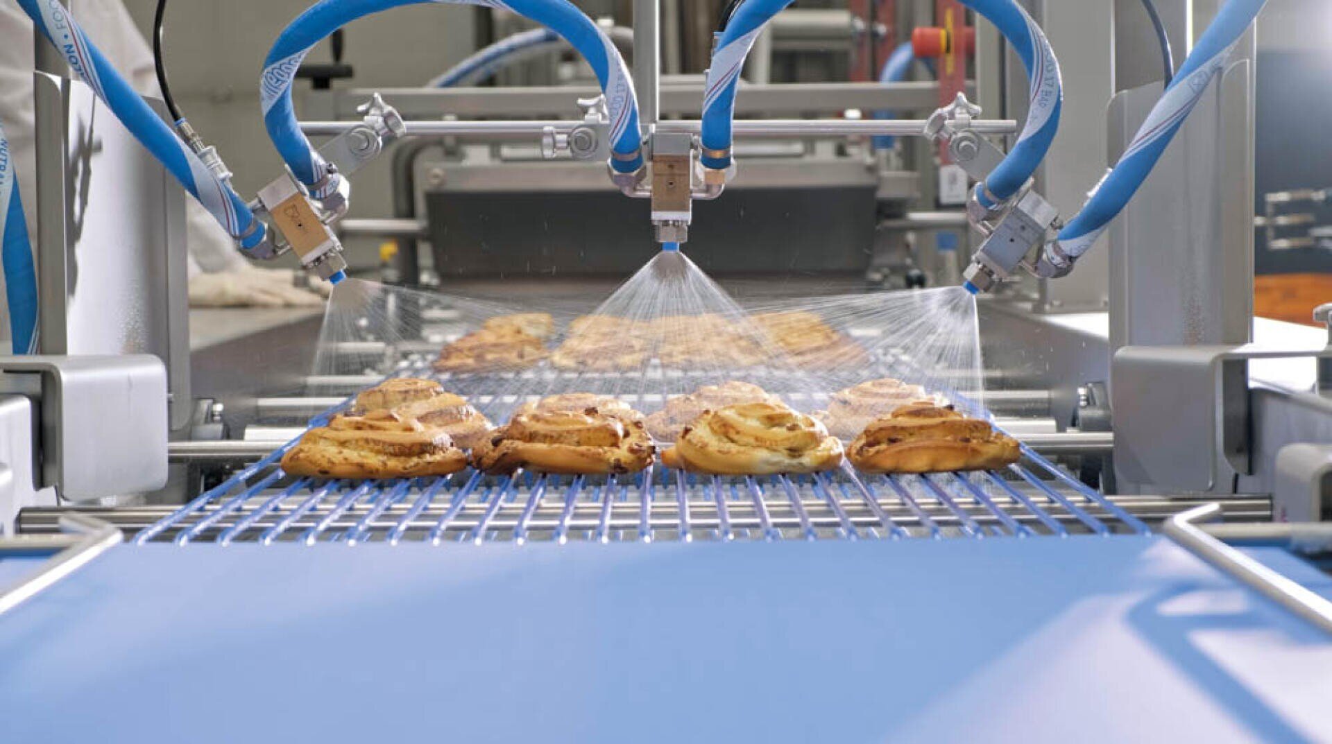 A row of pastries moves along a blue conveyor belt while multiple nozzles spray a mist over them in a food processing facility.