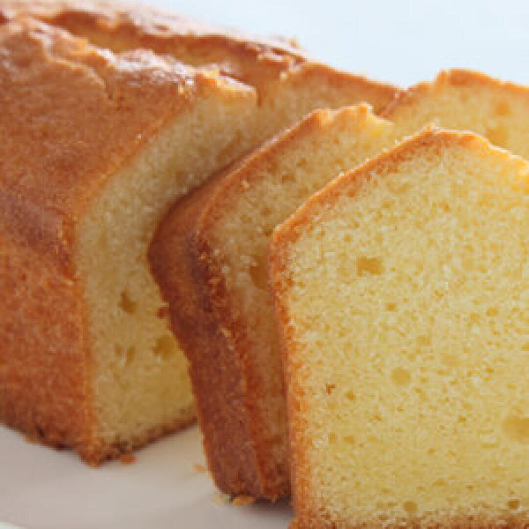 A close-up of a loaf of pound cake with a golden-brown crust, partially sliced to reveal its moist, light yellow interior. The cake slices are neatly arranged on a white surface.