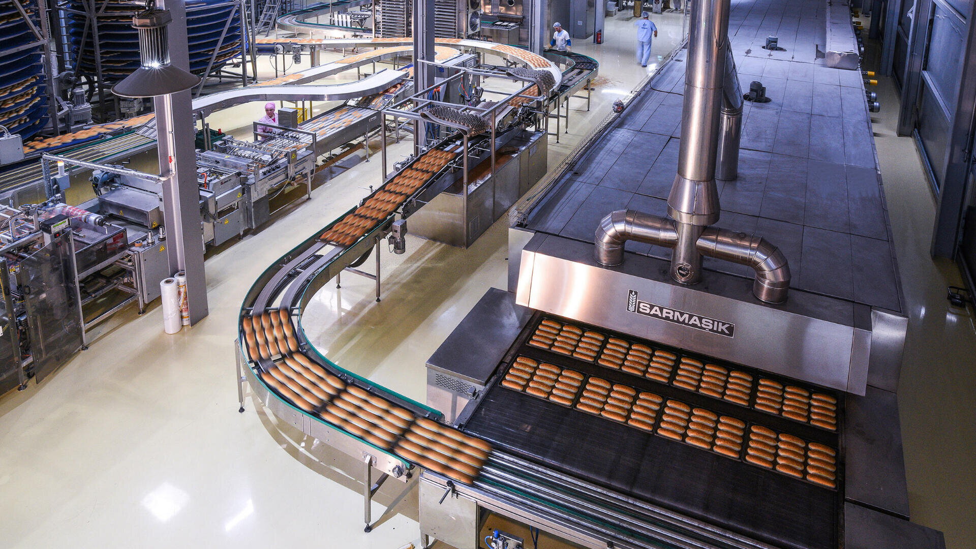 Förderbänder in einer Großbäckerei transportieren Reihen von frisch gebackenen Brotlaiben durch Maschinen, im Hintergrund sind Arbeiter in Schutzkleidung zu sehen.