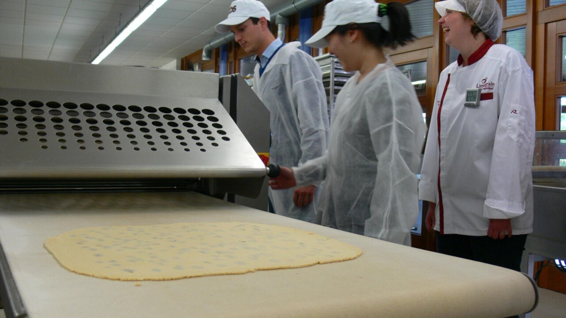 Three people in lab coats and caps stand next to an industrial dough roller machine in a bakery, watching as a sheet of dough moves along a conveyor belt.