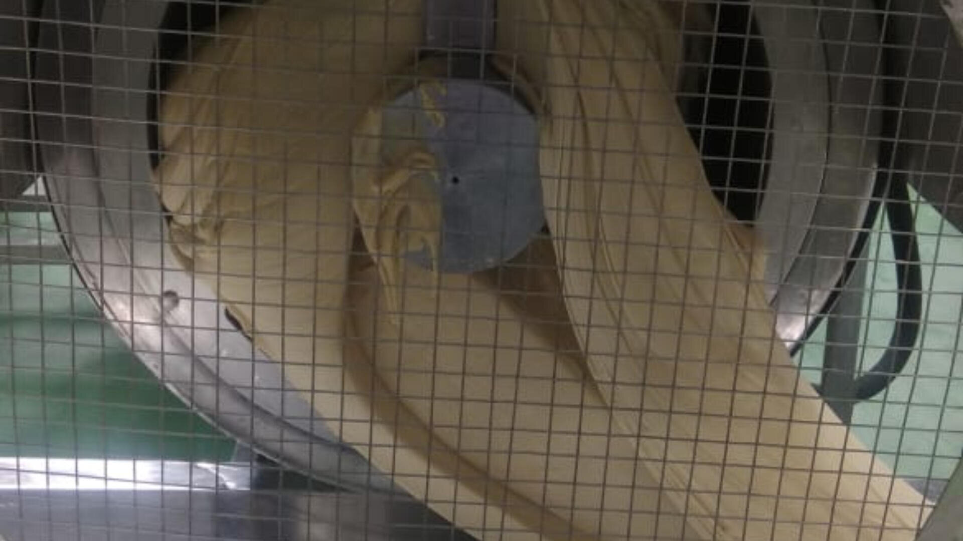 A close-up view of dough being kneaded in a large industrial mixer, seen through a metal grid for safety. The dough appears smooth and elastic as it wraps around the mixer’s central attachment.