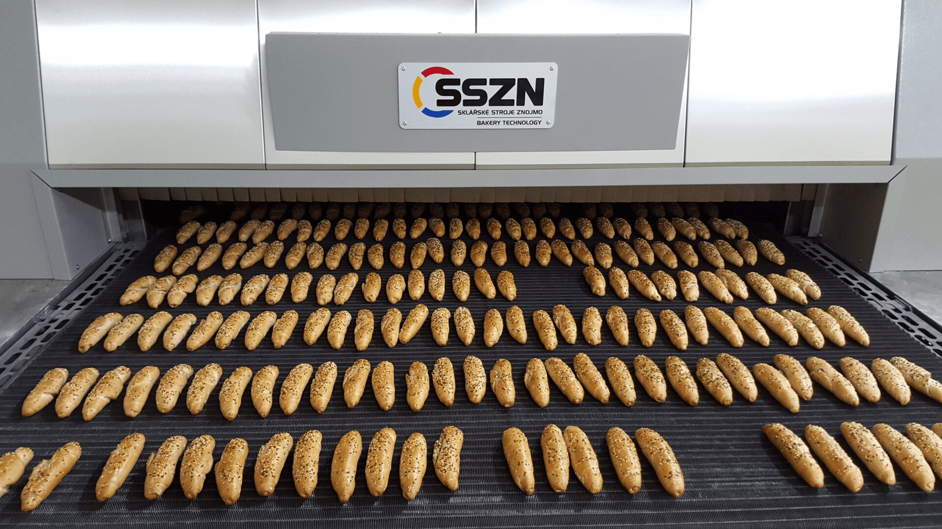 Rows of seeded baguettes on a conveyor belt coming out of a large industrial bread oven labeled SSZN.