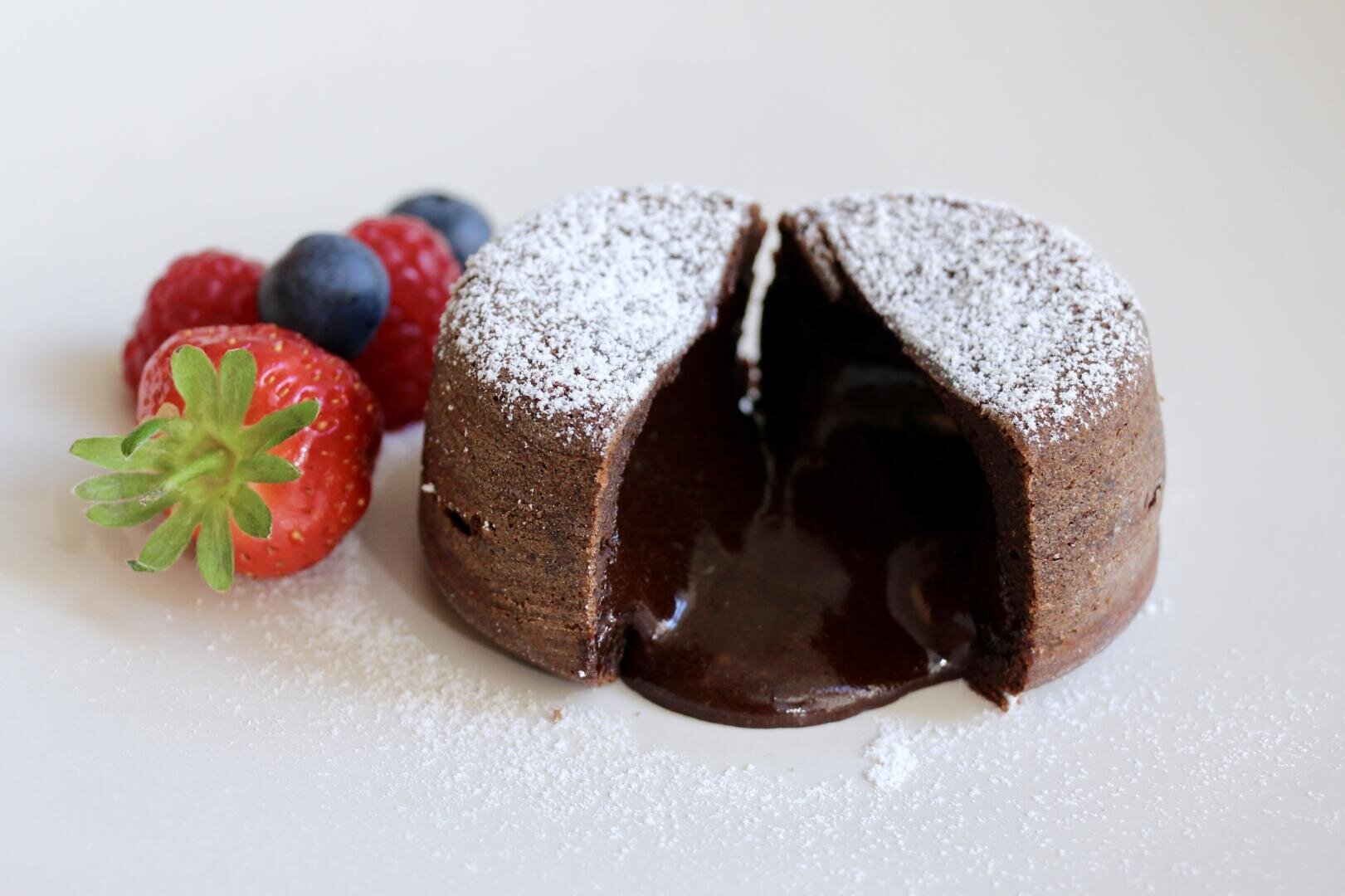 Ein Schokoladen-Lavakuchen mit herausquellender klebriger Füllung, bestäubt mit Puderzucker, wird auf einem weißen Teller mit frischen Erdbeeren, Blaubeeren und Himbeeren an der Seite serviert.
