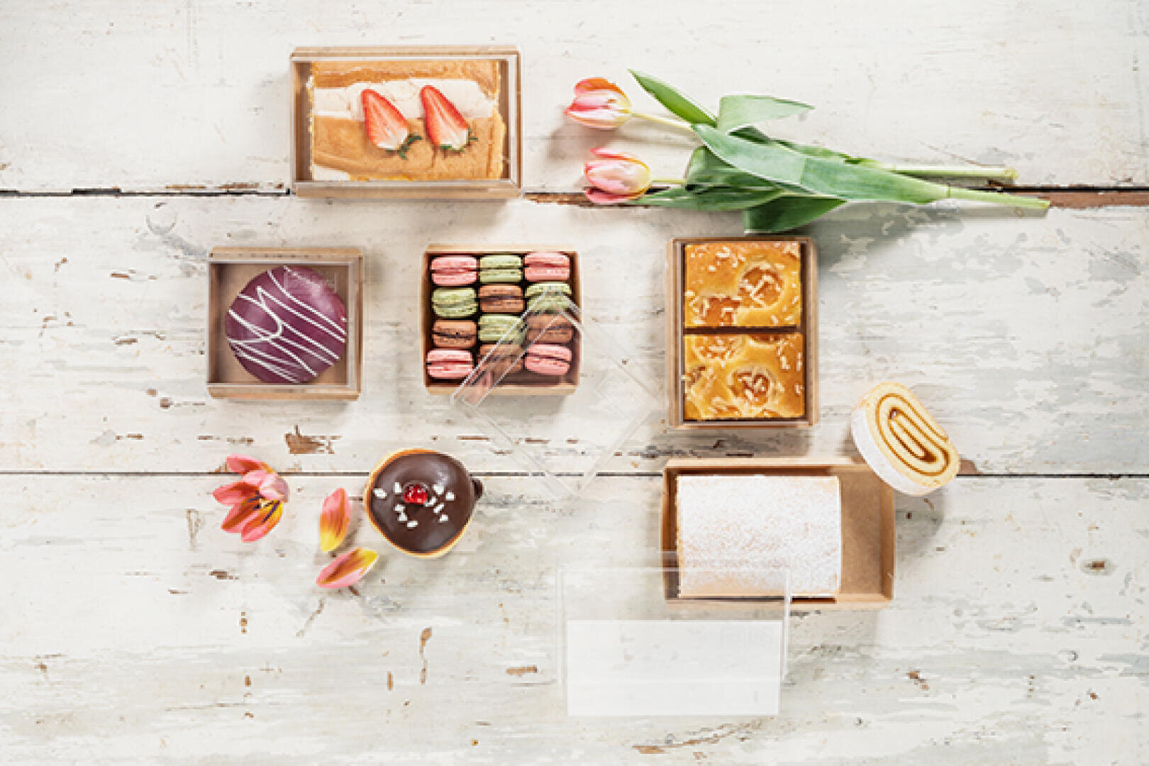 Ein Sortiment von Gebäck und Desserts in Schachteln, die auf einer rustikalen weißen Holzfläche mit rosa Tulpen und verstreuten Blütenblättern zur Dekoration angeordnet sind.