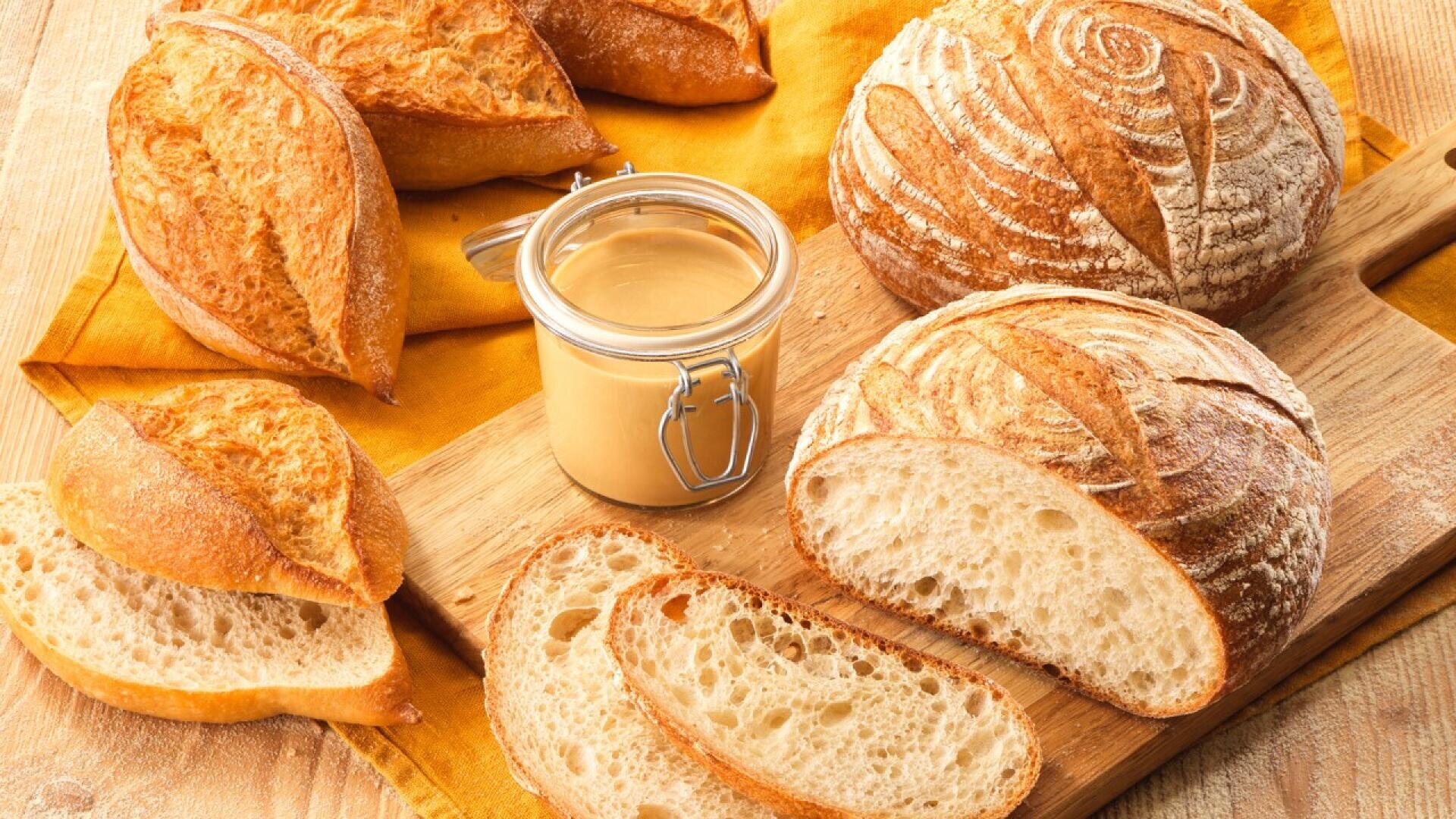 A wooden board with round loaves and rolls of crusty artisan bread, sliced pieces, and a small glass jar of creamy spread on a yellow napkin.