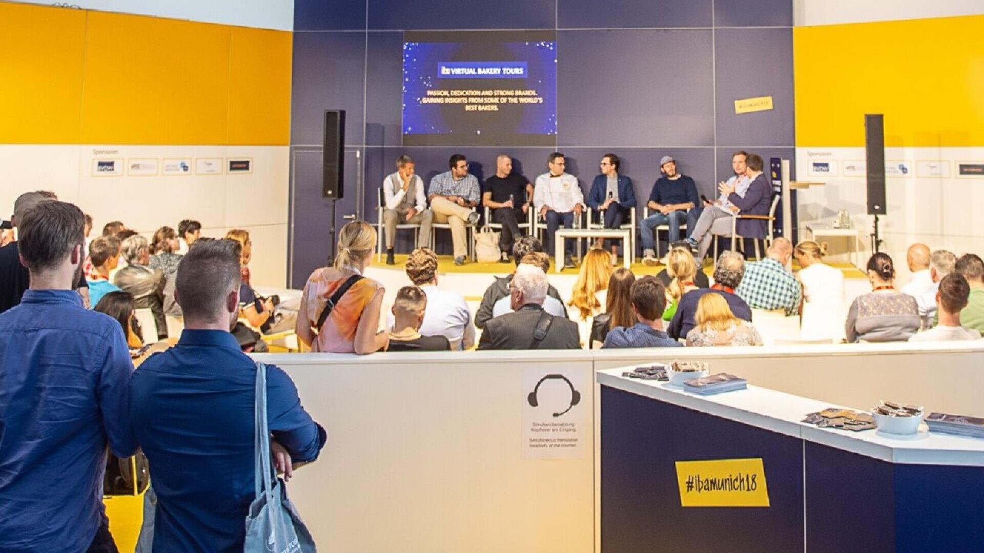 A group of seven people sits on a stage, speaking on a panel in front of an audience at an indoor event. Attendees are seated and standing, watching the discussion. Bright blue and yellow walls and a large display screen are visible.