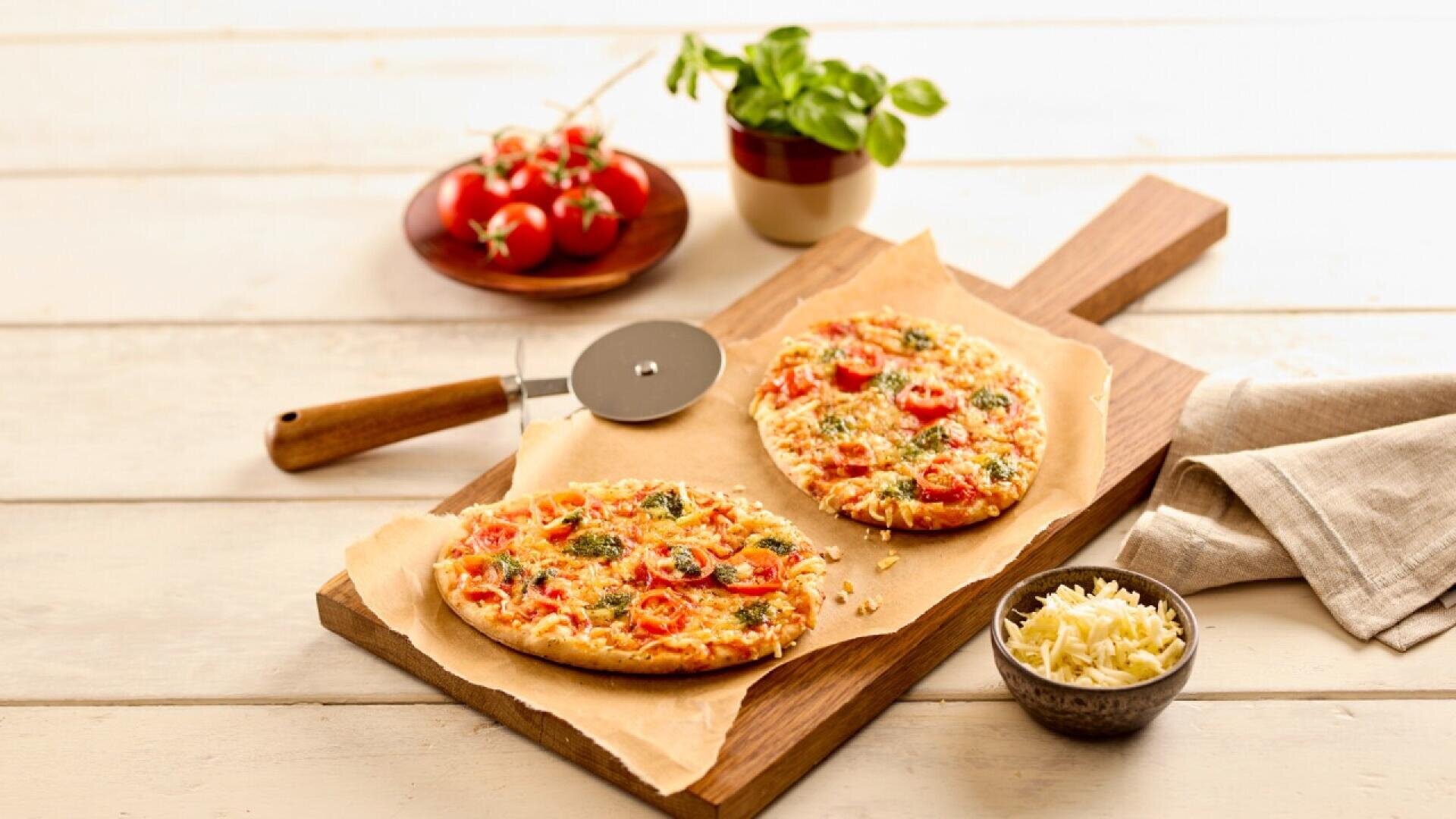 Two flatbread pizzas with cheese, cherry tomatoes, and herbs on parchment paper atop a wooden board. A pizza cutter, grated cheese, basil plant, and vine tomatoes are arranged nearby on a light wooden table.