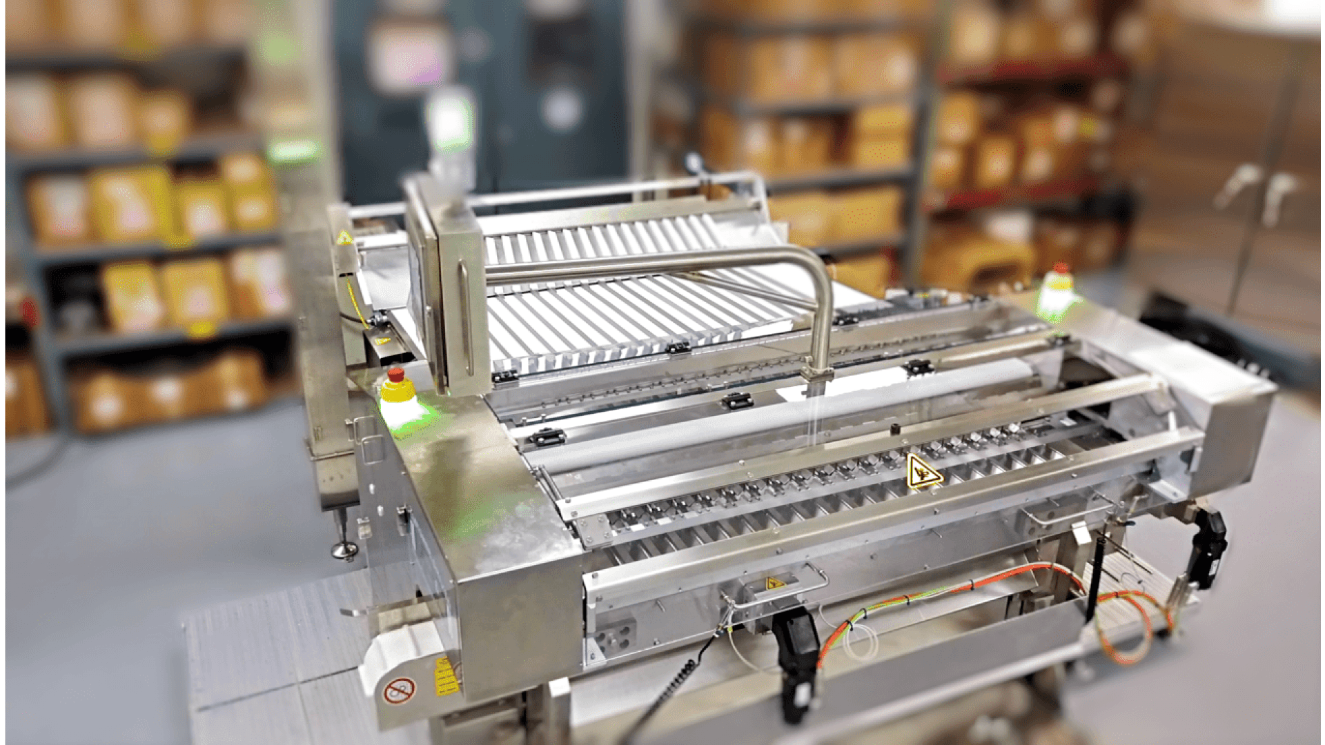 Ein Fördersystem aus rostfreiem Stahl in einem Lager, umgeben von Regalen mit braunen Kartons, mit Sicherheitsschildern, elektronischen Bauteilen und einer Rollenbahn für den Transport von Waren.