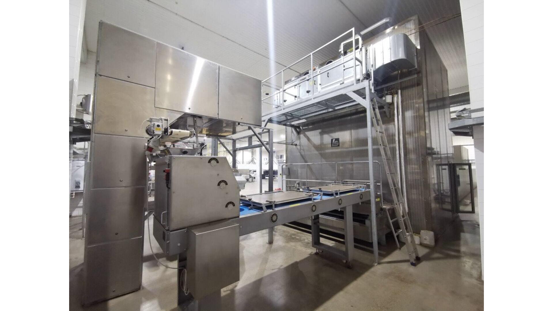 Eine große industrielle Lebensmittelverarbeitungsmaschine mit Metalloberflächen, einem Förderband und einer erhöhten Plattform befindet sich in einer hell erleuchteten, sauberen Fabrikumgebung. Eine Leiter führt zu einem erhöhten Laufsteg auf der rechten Seite.