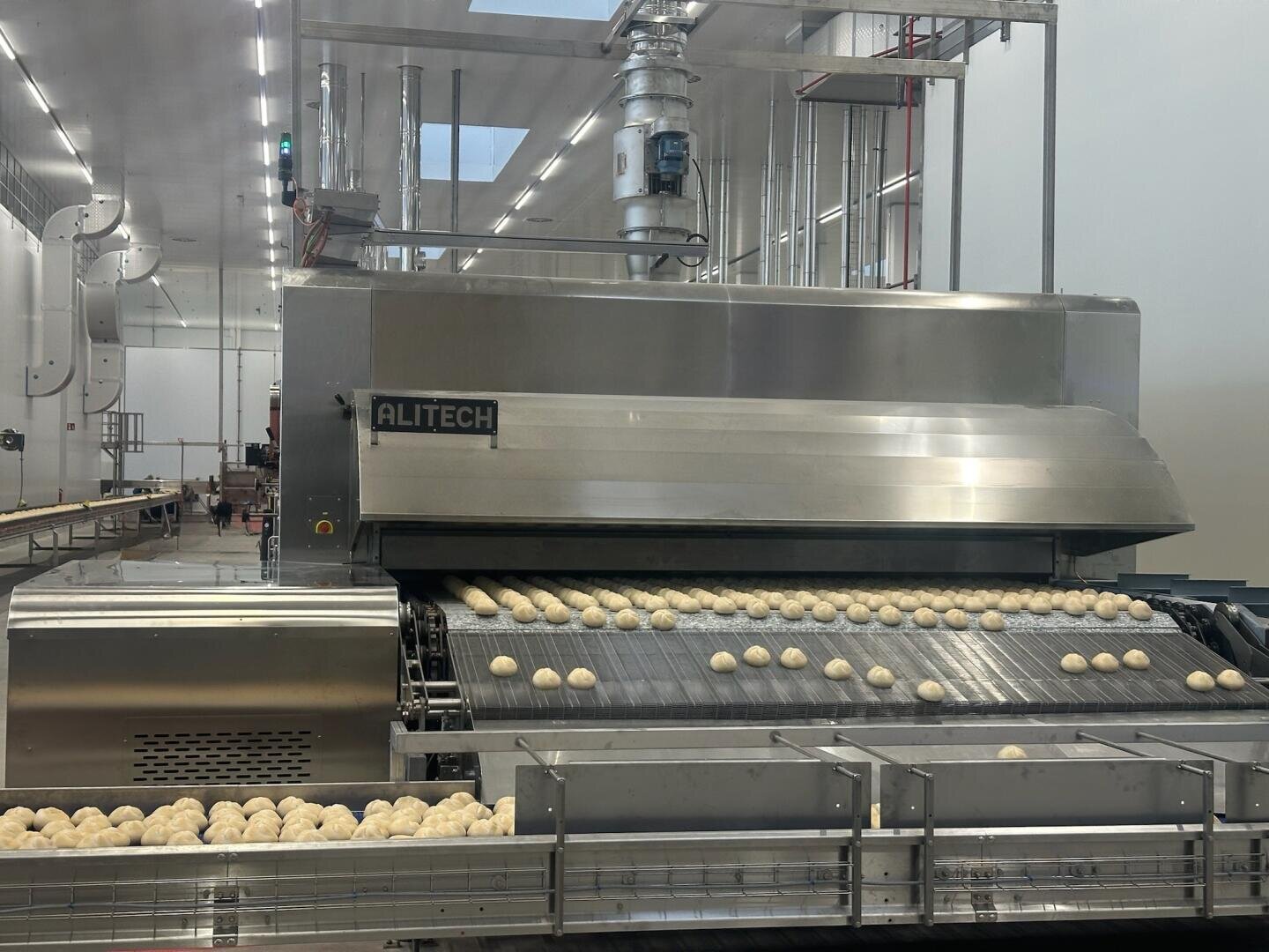Rows of dough balls move along a conveyor belt inside a modern industrial bakery, with large stainless steel machinery labeled ALITECH in a clean, well-lit facility.